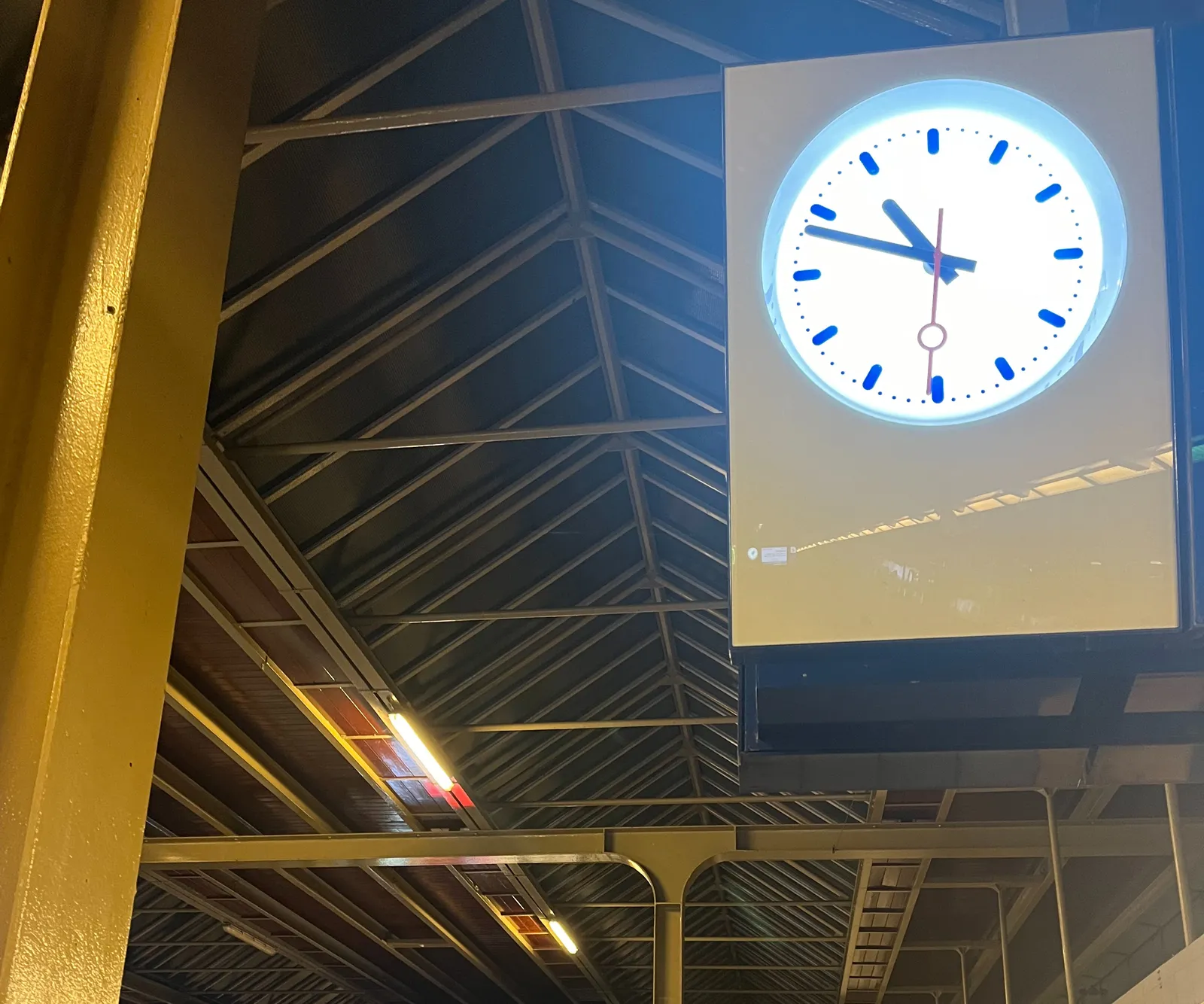 A metal construction bearing a roof, with a lit clock in the foreground. It points at 12 minutes to 11.
