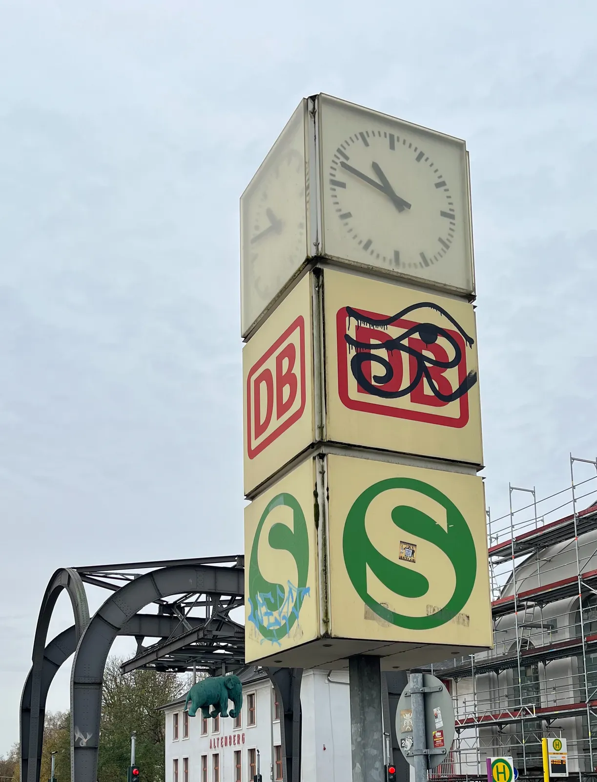 Large, in the foreground, a clock on a pole, with two large logos beneath it. The logos are from the german railways. Much more striking than this clock is the green elephant hanging from a heavy steel construction in the background. The clock points at 12 minutes to 11. 
