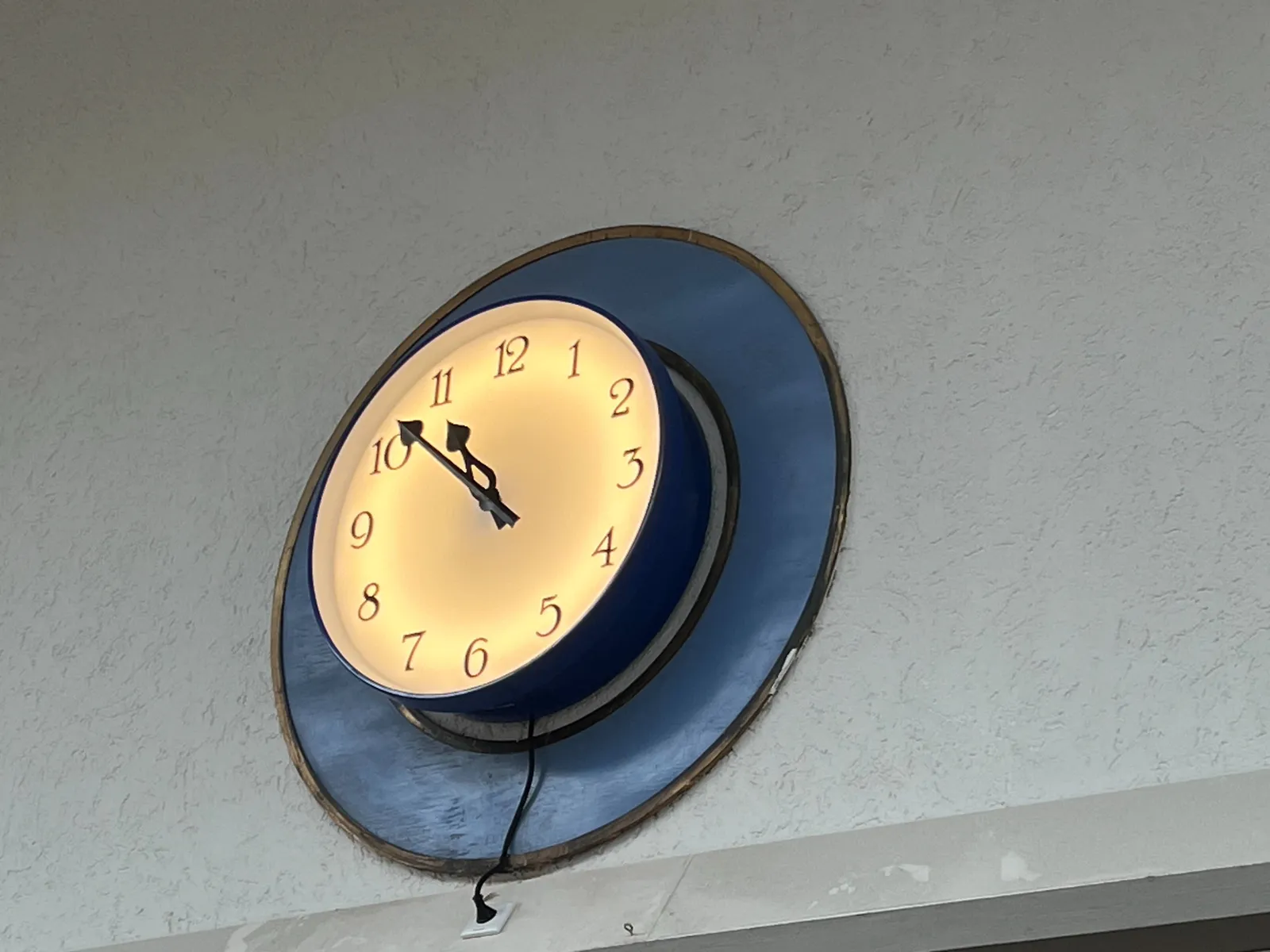 A clock that looks older that the plug that comes out of it. It’s placed inside a circular blue and copper frame, high on a wall. The clock points at 8 minutes to 11.