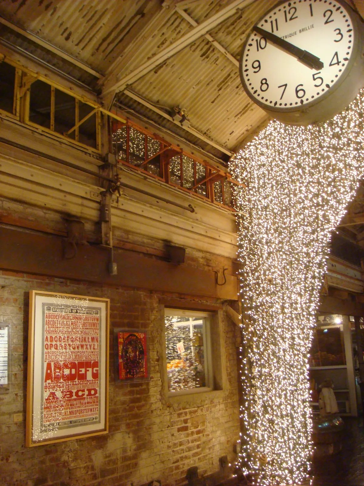 Inside an old industrial building. Brick walls, metal ceiling, and bright festive lights. There’s an old clock hanging from the ceiling. It points at 7 minutes to 11.