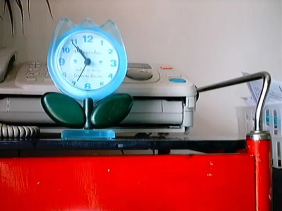 A clock in the shape of a flower, that stands in front of a fax machine, points at six minutes to 11.