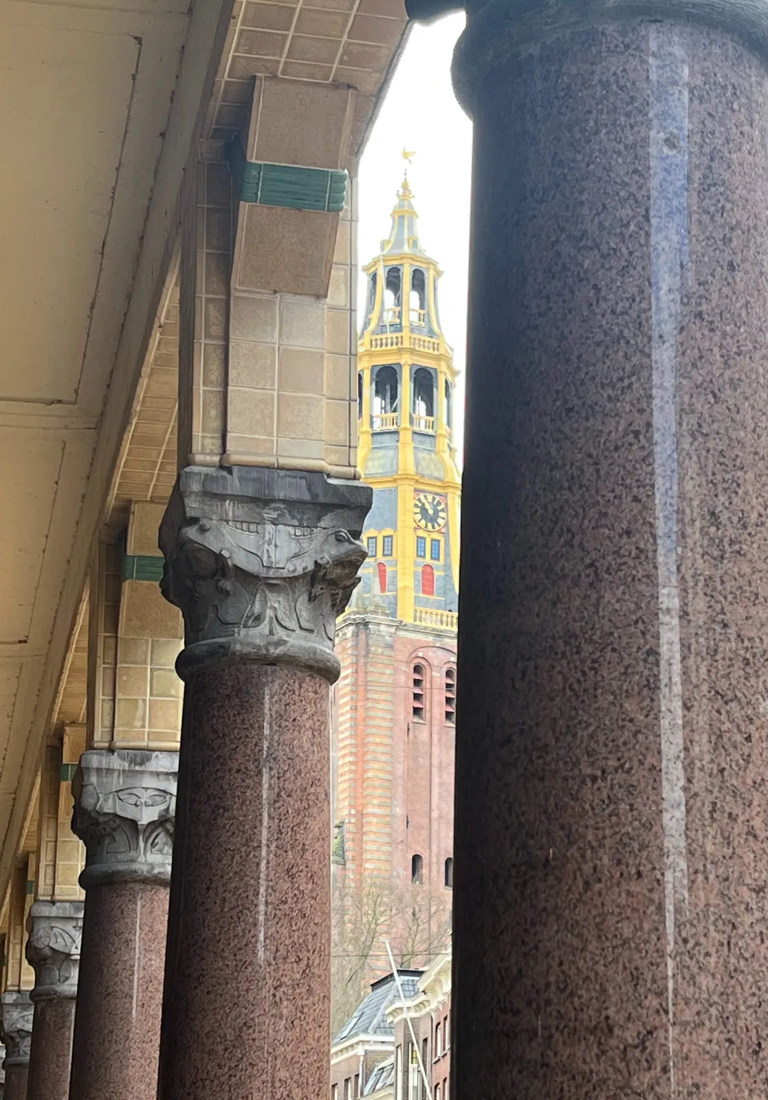 Peeking through the columns of a modern facade, clearly inspired by Korinthian columns. Between these columns you can see an old church tower with a clock on it. It points at 5 minutes to 11.