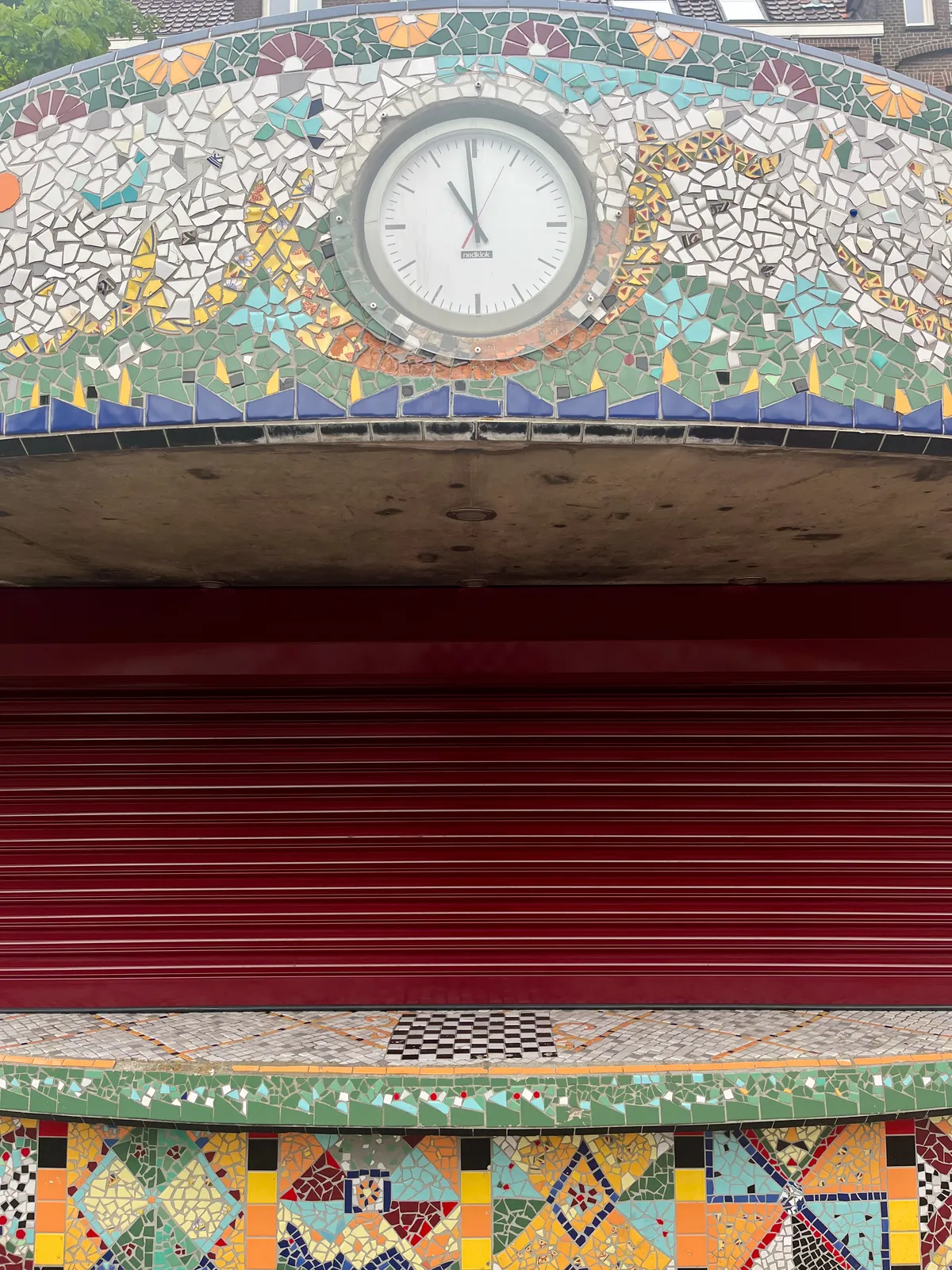 A clock built into some sort of a concrete kiosk decorated with a colourful mosaic. It points at one minute to 11.
