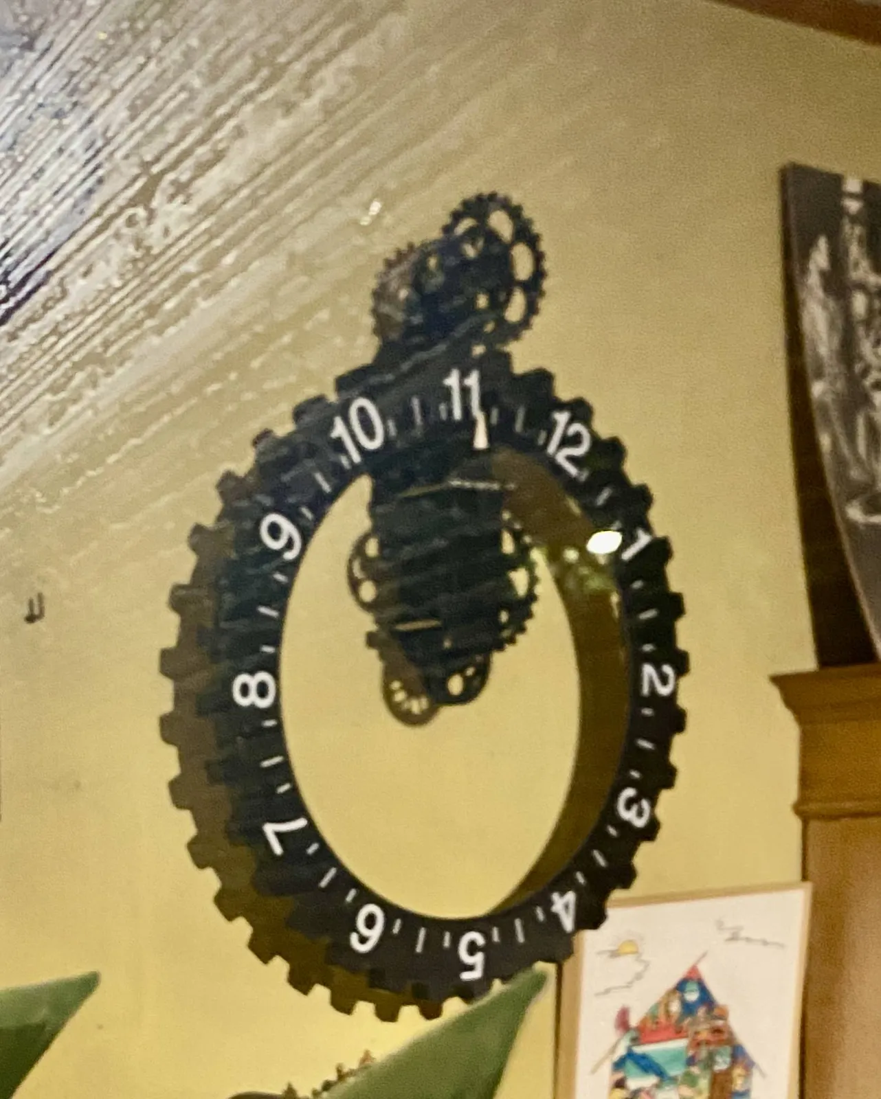 A black wall clock without hands. It’s made from a ring with the numbers 1 to 12 on it. It’s this ring that turns past a simple white triangular indicator. The mechanics of this clock are visible.  It points at 11 o’clock.