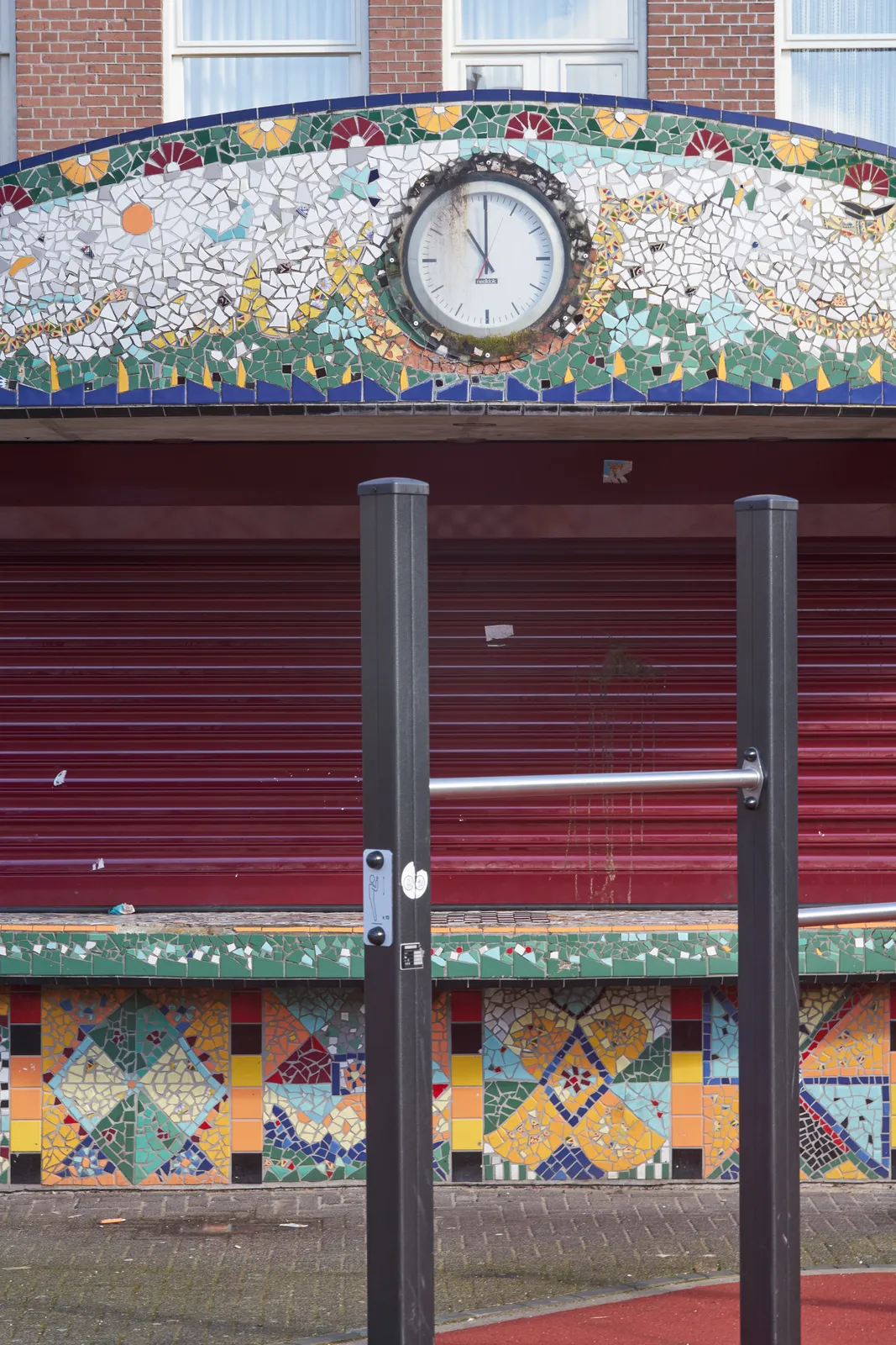 A mosaic on a kiosk in a playground. There’s a clock in this mosaic and it points at 11 o’clock.