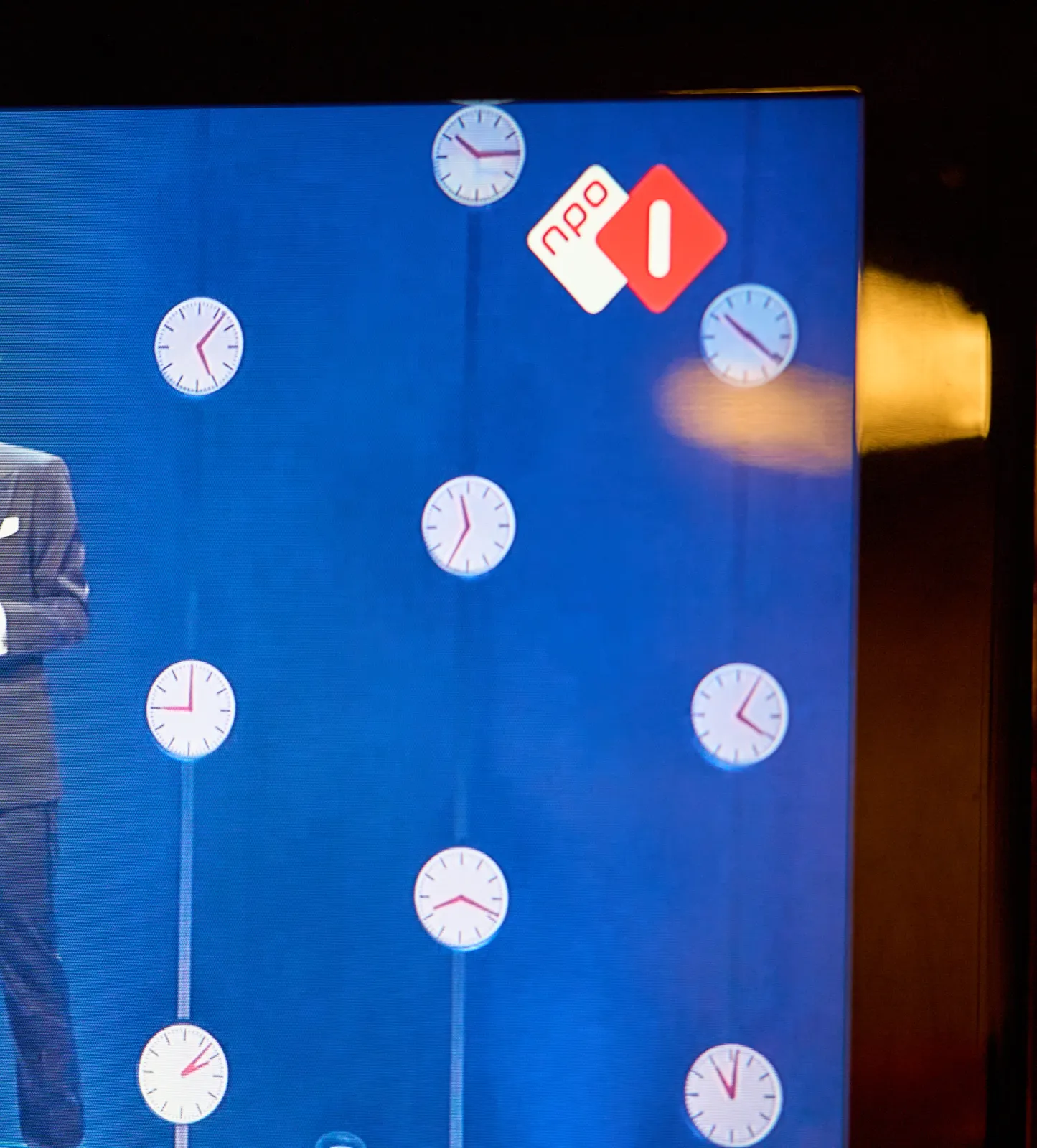 A picture taken from a television. You can see someone in a black suit standing, and behind him there’s a set made from lots of clocks. Nine clocks are visible. They point at a quarter past ten, six minutes past five, 21 minutes past ten, 25 minutes to 12, nine o’clock, five past 4, 18 minutes past 8, 7 minutes past 2, and the last one points at one minute past 11. One of these clocks points at the right time.