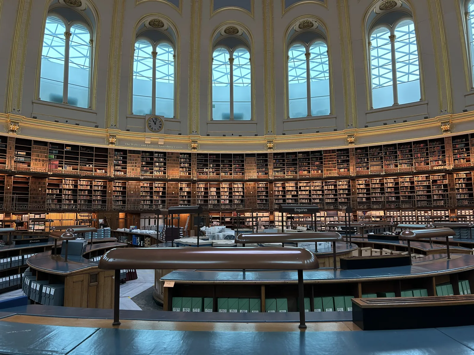 A library in a large, high oval building. The bookcase fills the lower part of the wall, large windows above it. There’s a clock placed above the bookcase, and above the label that says The Walter and Leonore Annenberg Centre. This clock points at two minutes past eleven. 
