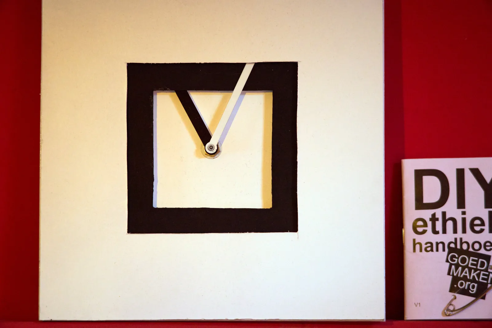A square, blanc and white clock with hands that seem to grow. It’s an abstract, a bit arty thing. Next to it stands a copy of the DIY ethics handbook. The clock points at 4 minutes past 11.