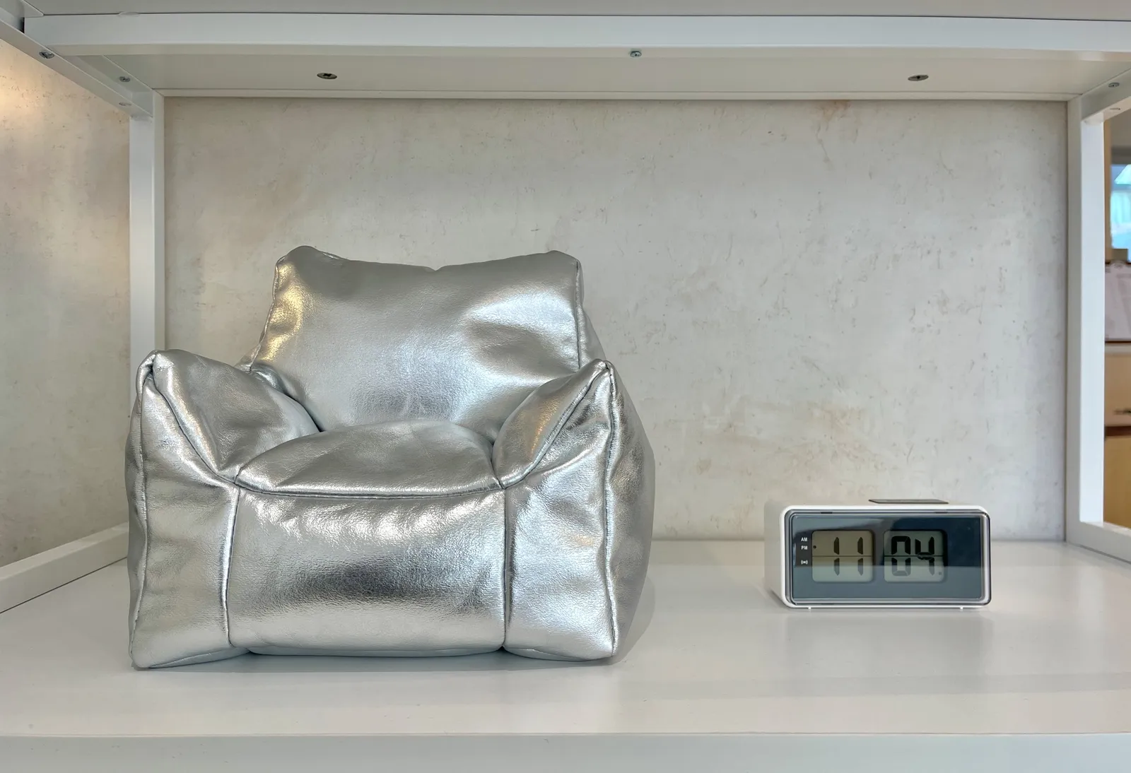 A silvery shiny model of a designer chair. Next to it there’s a digital clock, that pretends to be a flip clock, and pretends to be a segmented clock at the same time. It shows the time as 11:04.