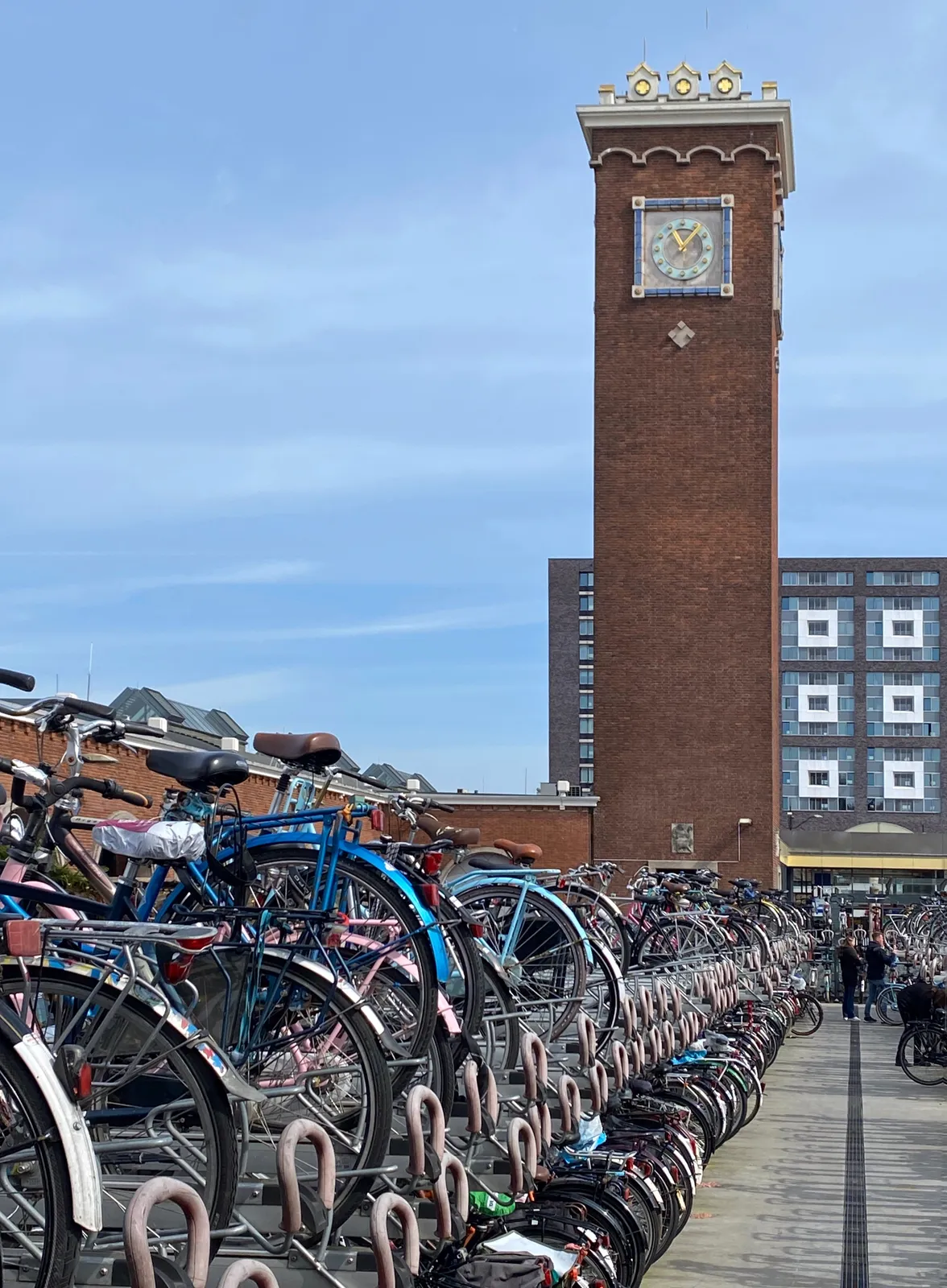 A double layered, very long bicycle storage construction, filled with bicycles. Behind it is a tower with a clock on it. The clock points at 6 minutes past 11.