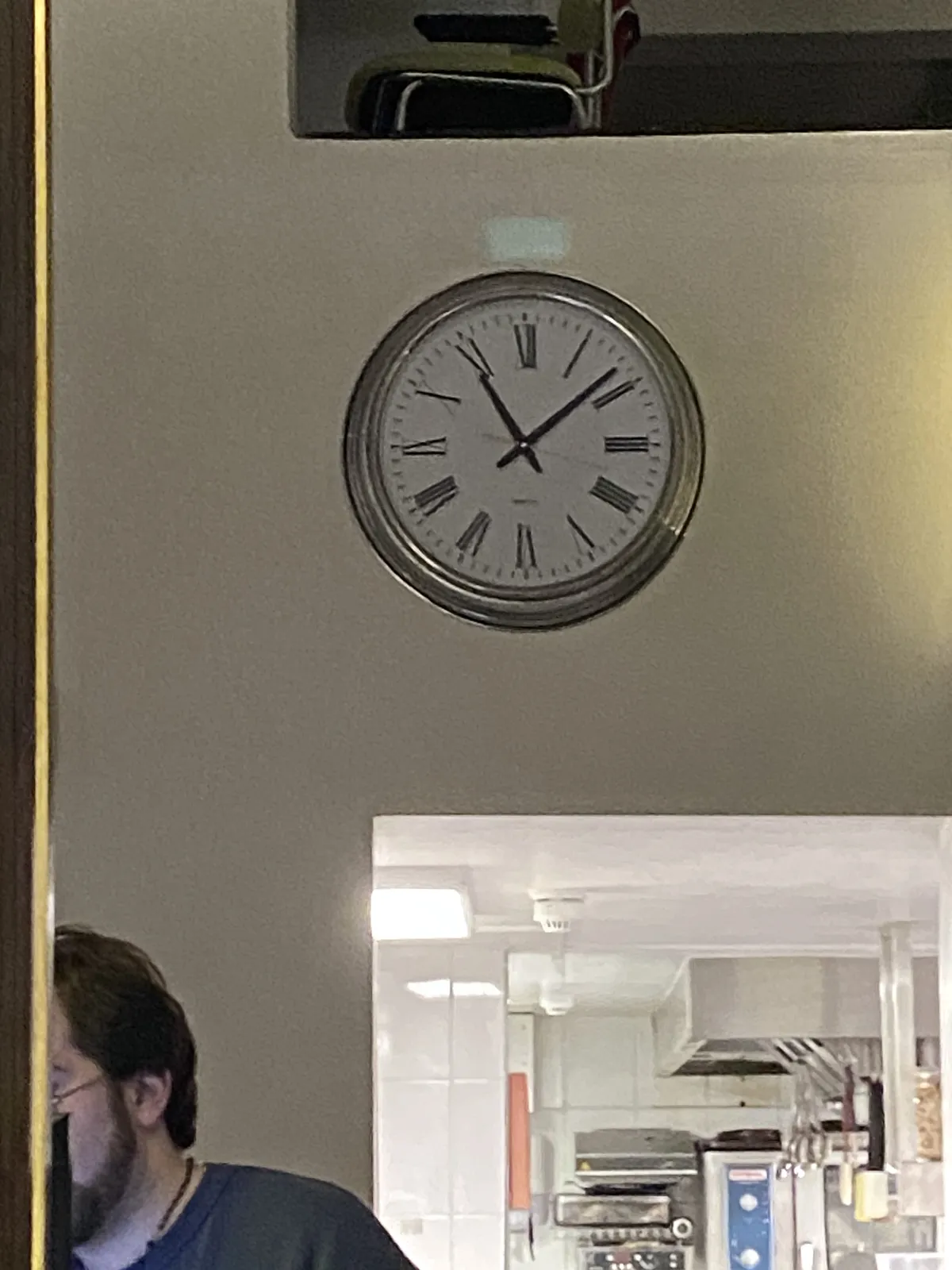 An interesting picture. There’s a person in this picture, but they’re cut off by a pole, and by the frame of the picture. You can look through a door into what seems to be a professional kitchen. And upstairs you can look through a window. There’s a bauhaus type chair there. There’s also a clock on the wall. It points at 8 past 11.