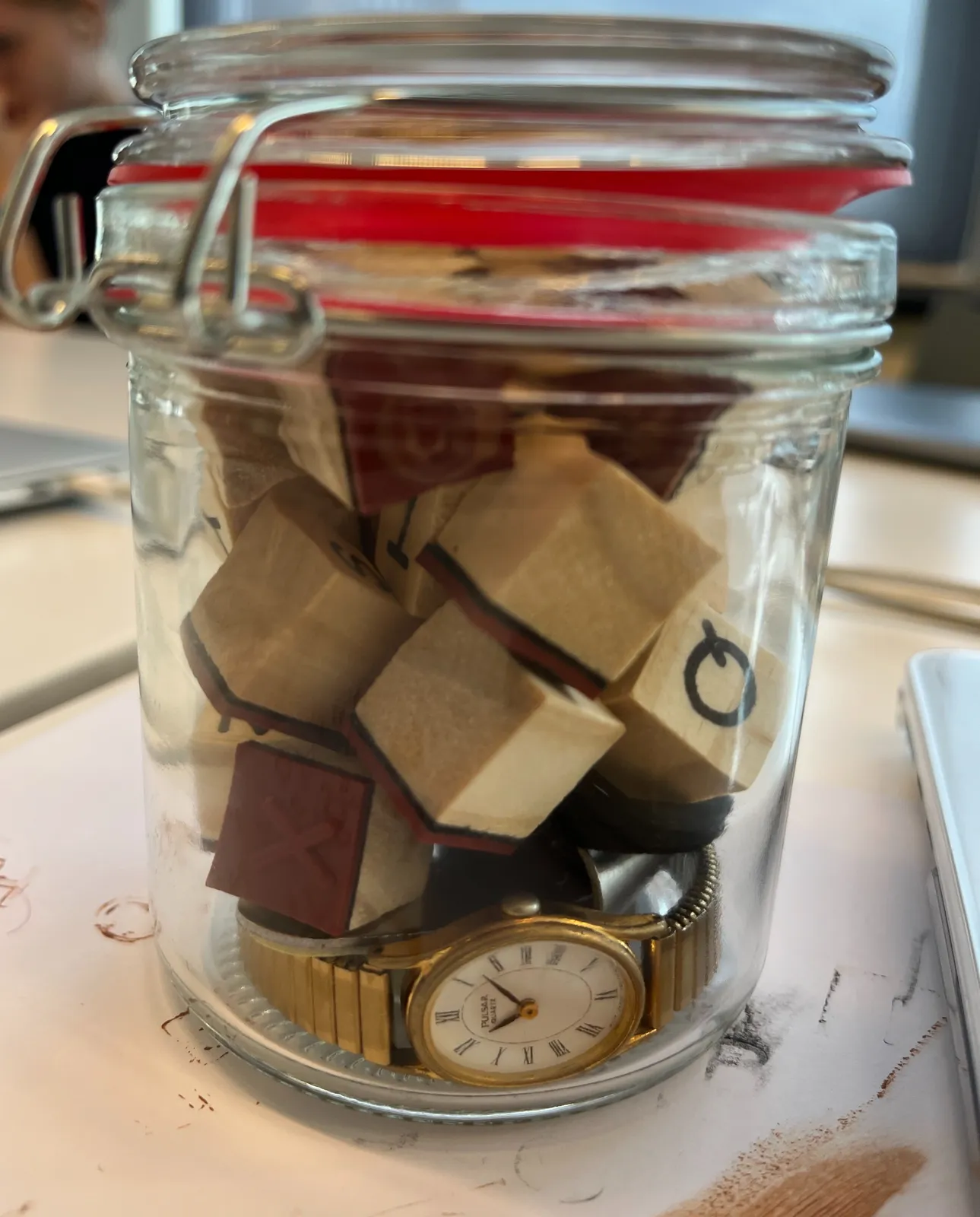 A glass jar filled with little wooden cubes, and a watch. The watch points at 8 minutes past 11.
