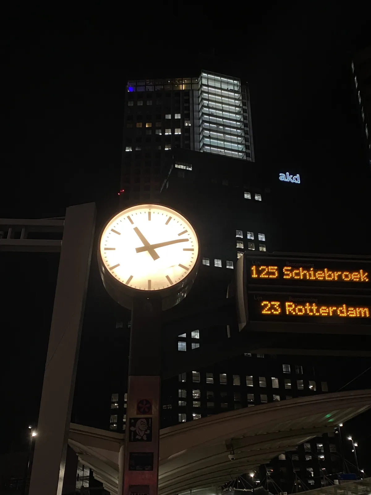 It’s dark outside. A few very high appartment buildings, or offices in the background. In the front there’s a lit bus stop sign. It says 125 Schiebroek and 23 Rotterdam. The lit clock next to it points at 12 past 11.