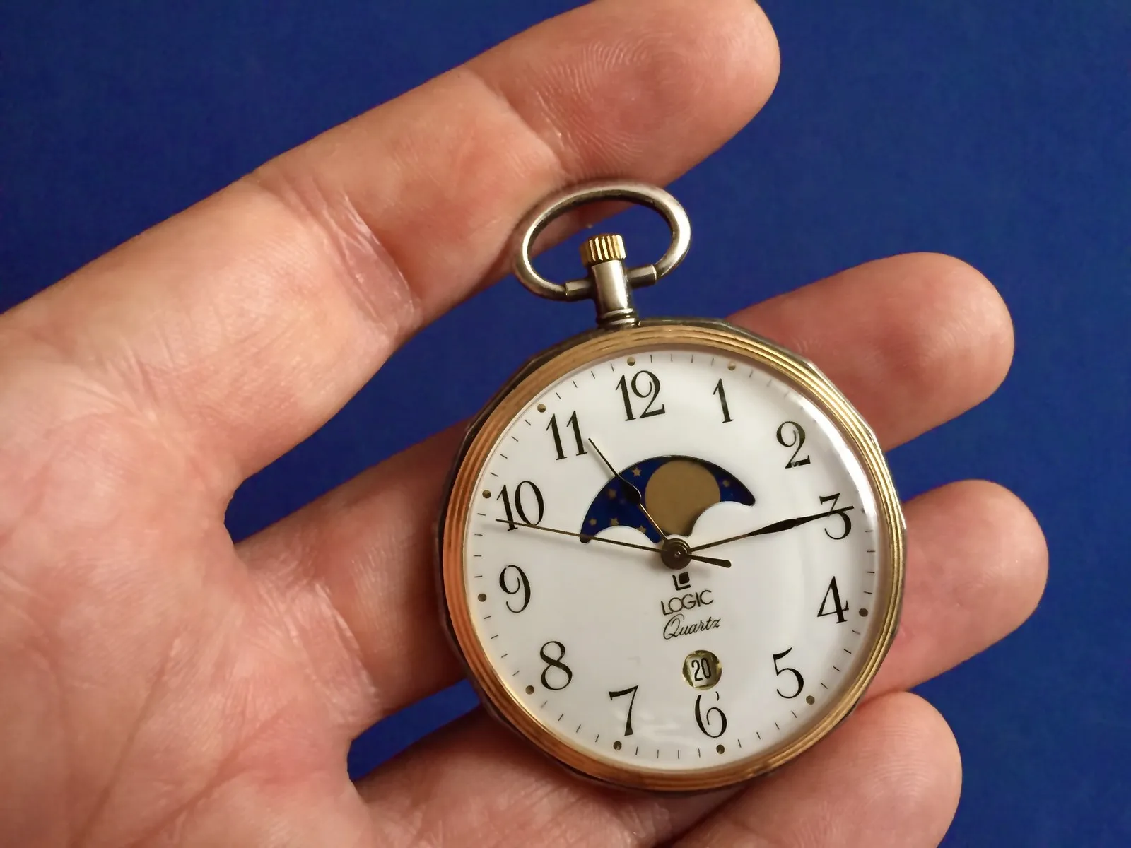 Someone holds a quartz pocket watch in their hand. It points at 14 minutes past eleven.
