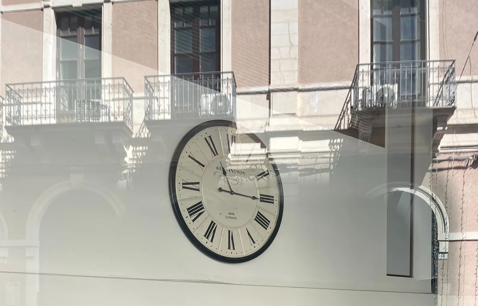 The reflection of a classical italian facade with balconies in a window. Behind the window there’s a clock that points at a quarter past 11.