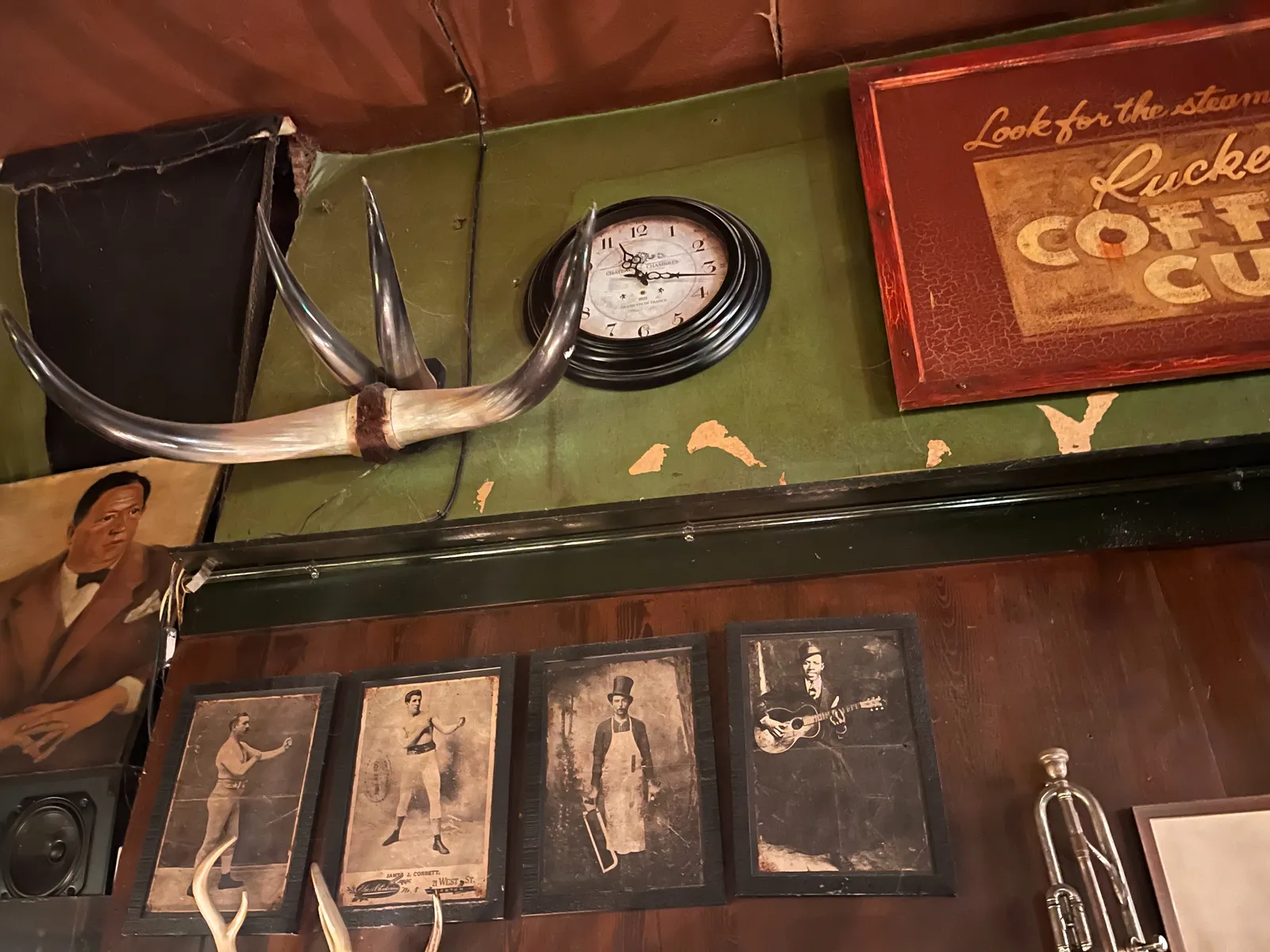 A wall with old black and white pictures of boxers, a butcher, and a blues guitarist. A part of a trumpet and part of an old billboard can be seen as well. There’s also a wall clock, partially hidden behind some very large cow horns. The clock points at 16 minutes past 11.