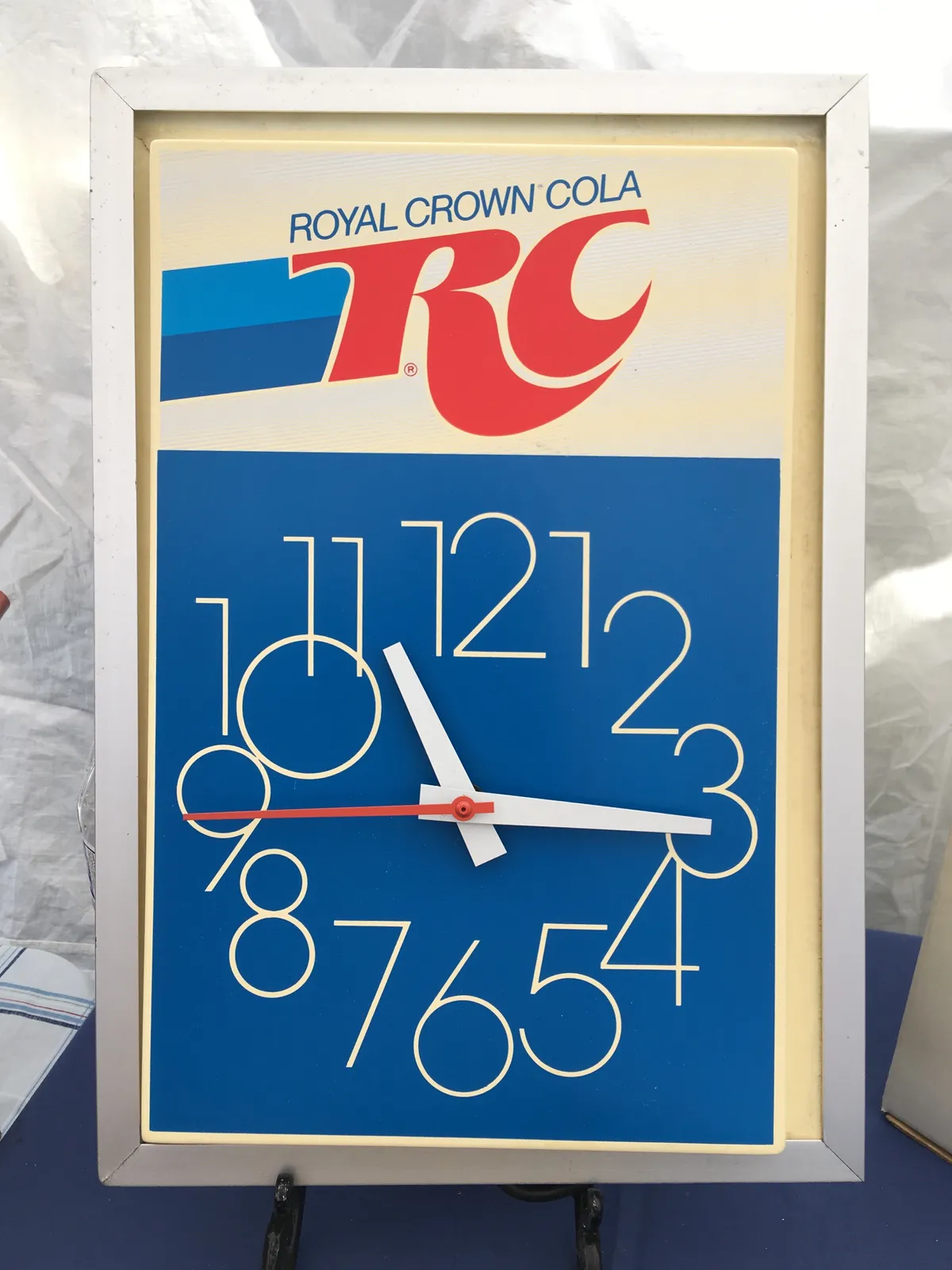 An advertisement for RC, Royal Crown Cola. Below it is a blue clock with numbers that are so large that some of them overlap. This clock points at 16 minutes past 11.
