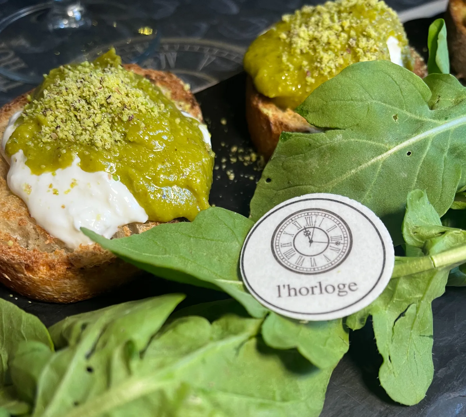 Two bruschettas with ricotta and pistachio paste on a plate. There are also large ruccola leaves on the plate. On the leaves there’s a round piece of paper with a clock printed on it that points at 17 minutes past 11. The word l’horloge is printed below it.