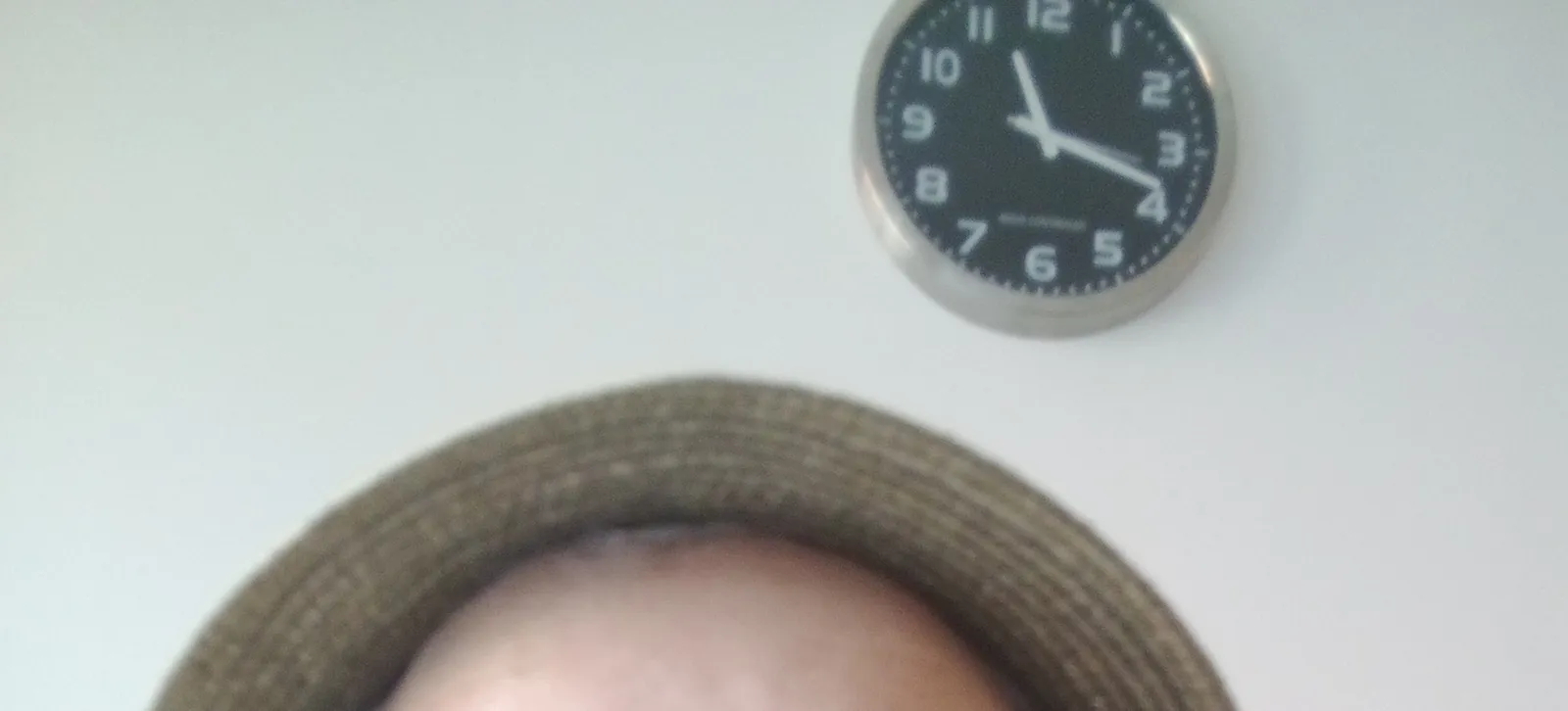 The top of someone’s head, with a hat on top. Behind it there’s a clock that points at 18 minutes past eleven.