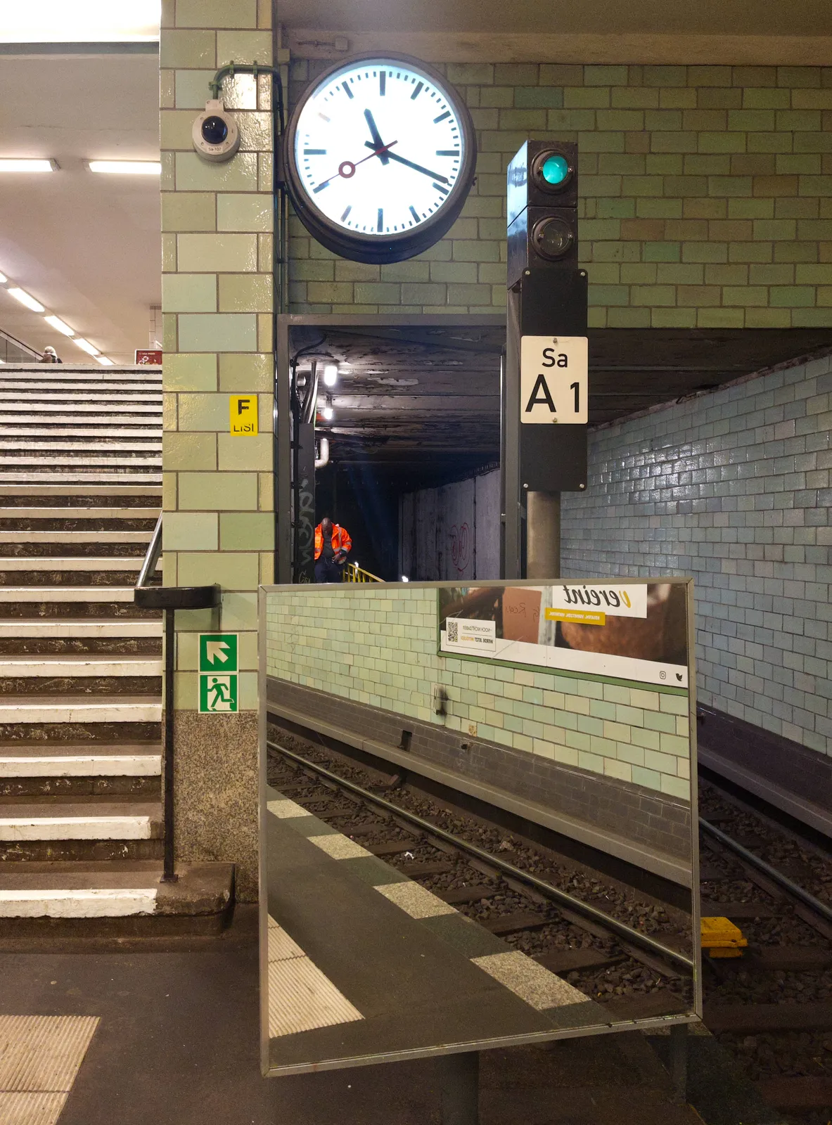 On the left there are stairs, going up. On the right there’s a tunnel. At the right bottom of the picture there’s a large square mirror, in which some train tracks can be seen. So it’s a train, or metro tunnel. Above this mirror there’s a clock which points at 19 minutes past 11.