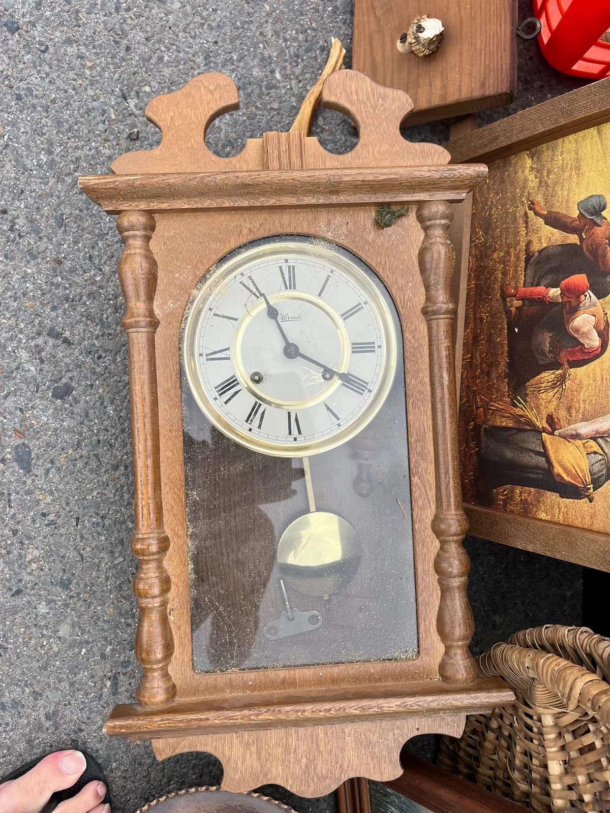 A painting, and a wooden clock for sale, both laying on the ground. The clock points at 20 past 11. The toes of the photographer can be seen, in their flipflops. 
