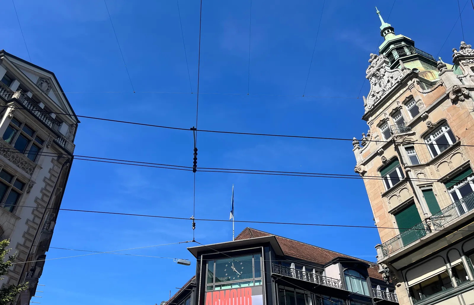 Looking at a clear blue sky and some buildings. On one of these buildings there’s a clock. It points at 22 minutes past 11.
