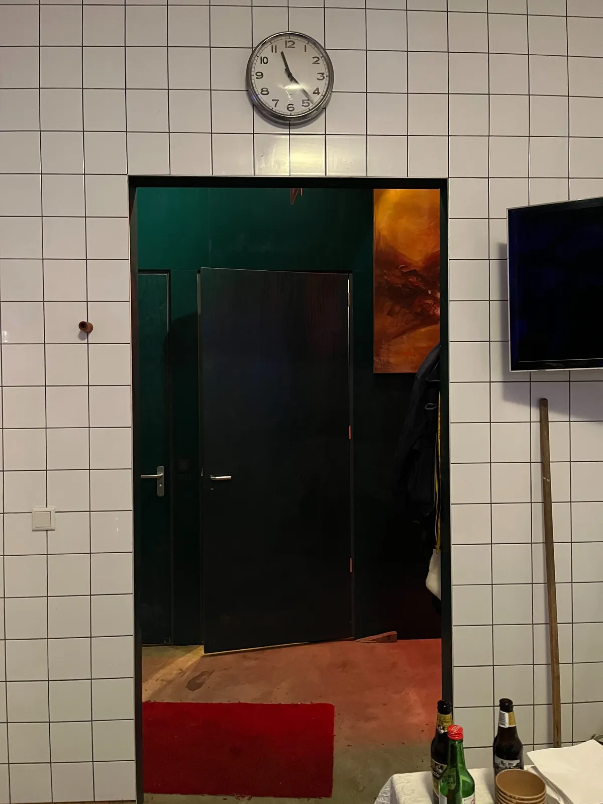 Peeking through a door, at a dark green wall, with two doors in it. There’s also an expressive painting on this wall. The room the photographer is in, is completely covered in white tiles. Above the door is a simple clock. It points at 23 minutes past 11