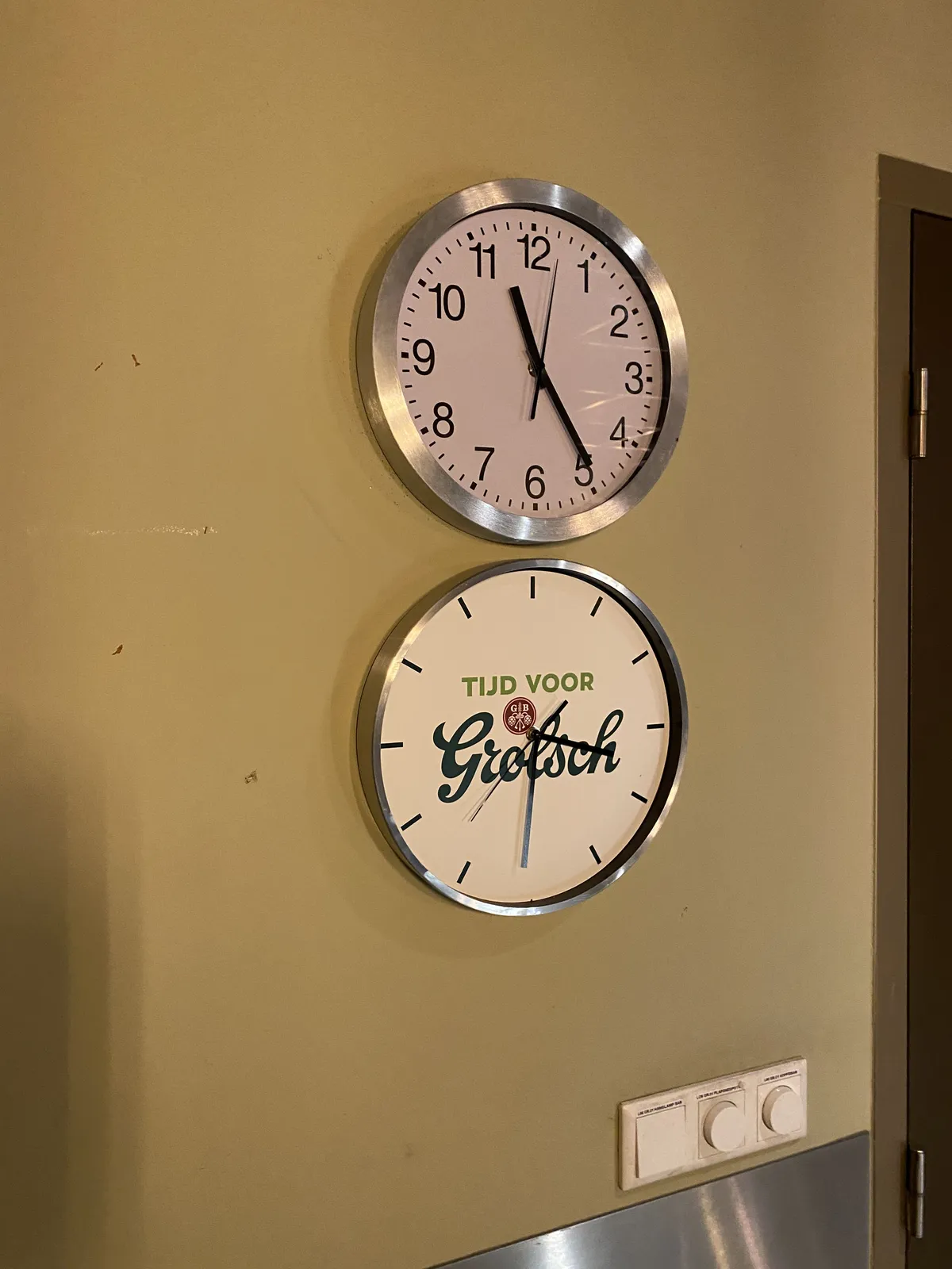 There are two wall clocks, one hanging right above the other. The top one, a simple, modern minimalist clock, points at 24 minutes past 11. The other one, right below it, that says it is time for Grolsch, points at 31 minutes past 3.