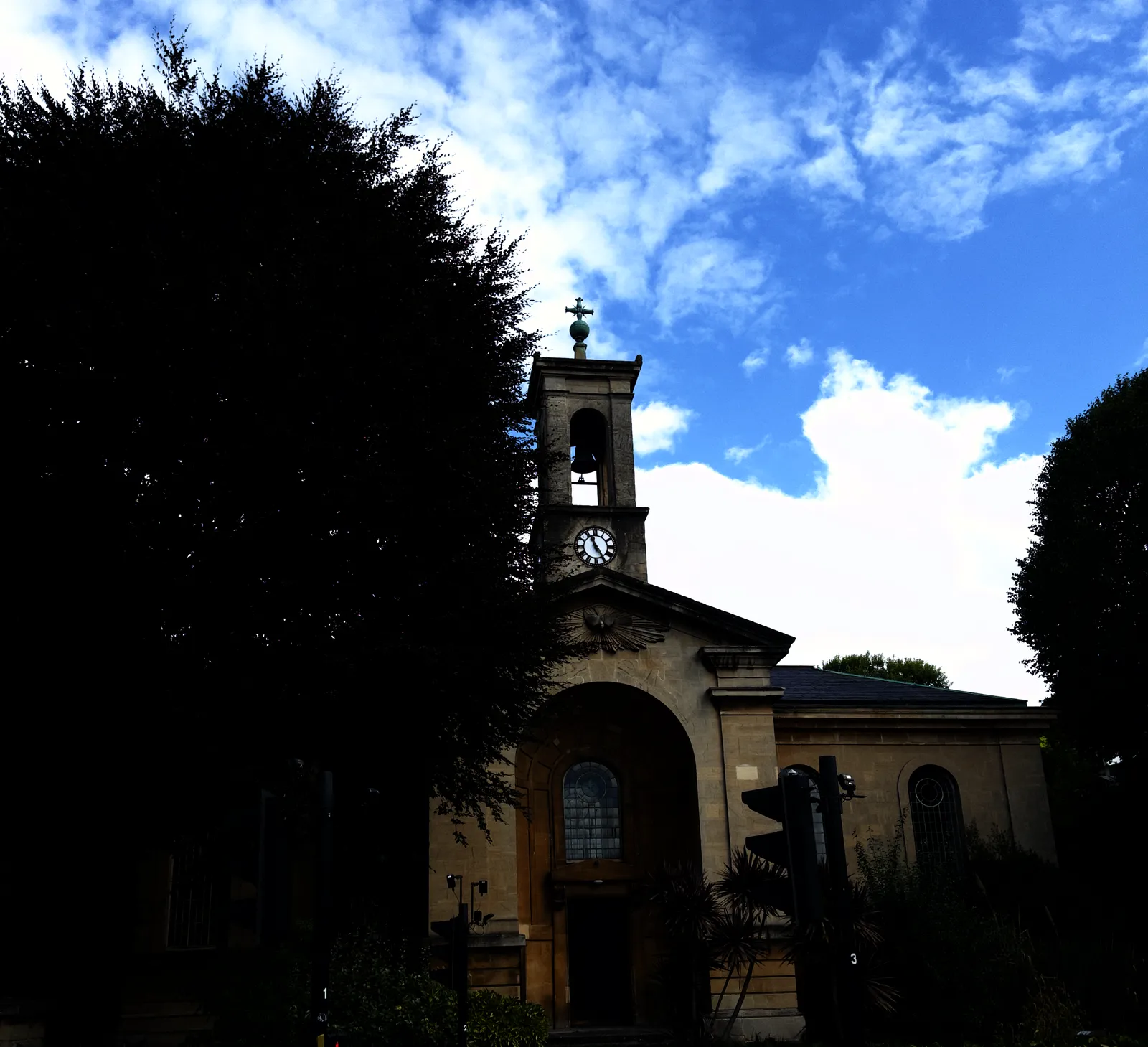 A not too large church, half hidden behind a large tree. The clock on the church tower points at 25 minutes past eleven.