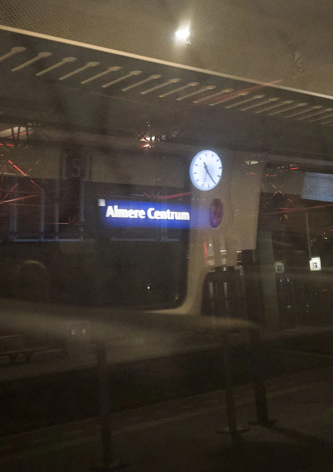 Peeking out the window of a train. It’s dark outside. The station sign of Almere Centrum is lit. A clock is lit as well. It points at 25 past 11.