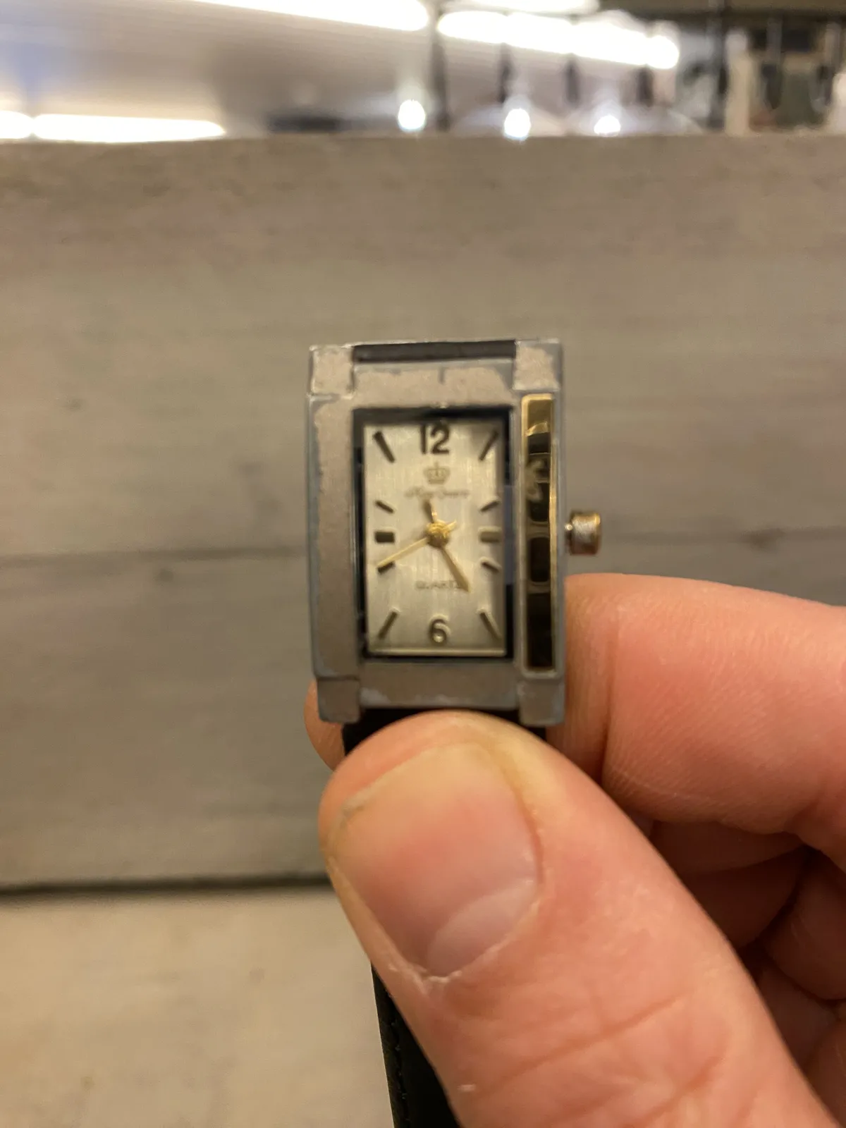 A small rectangular wrist watch. It points at 25 minutes past 11. 
