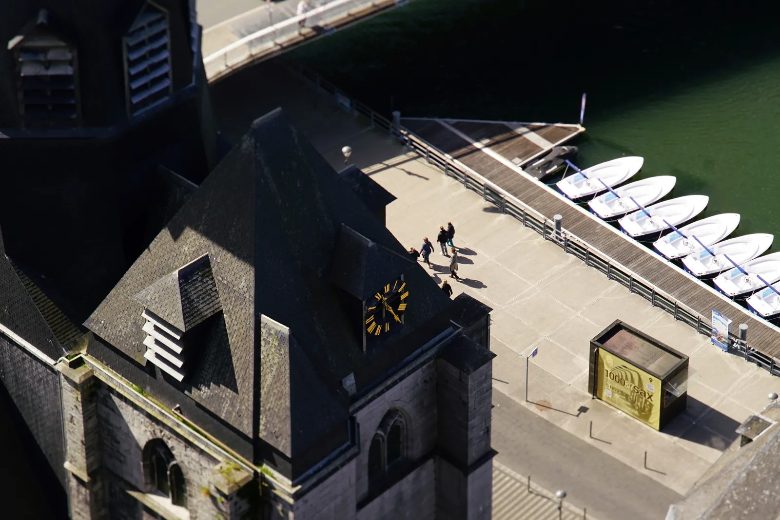 Looking down from a pretty high place upon a harbor with some small boats, some people walking and an old building with a clock. It points at 26 minutes past 11.