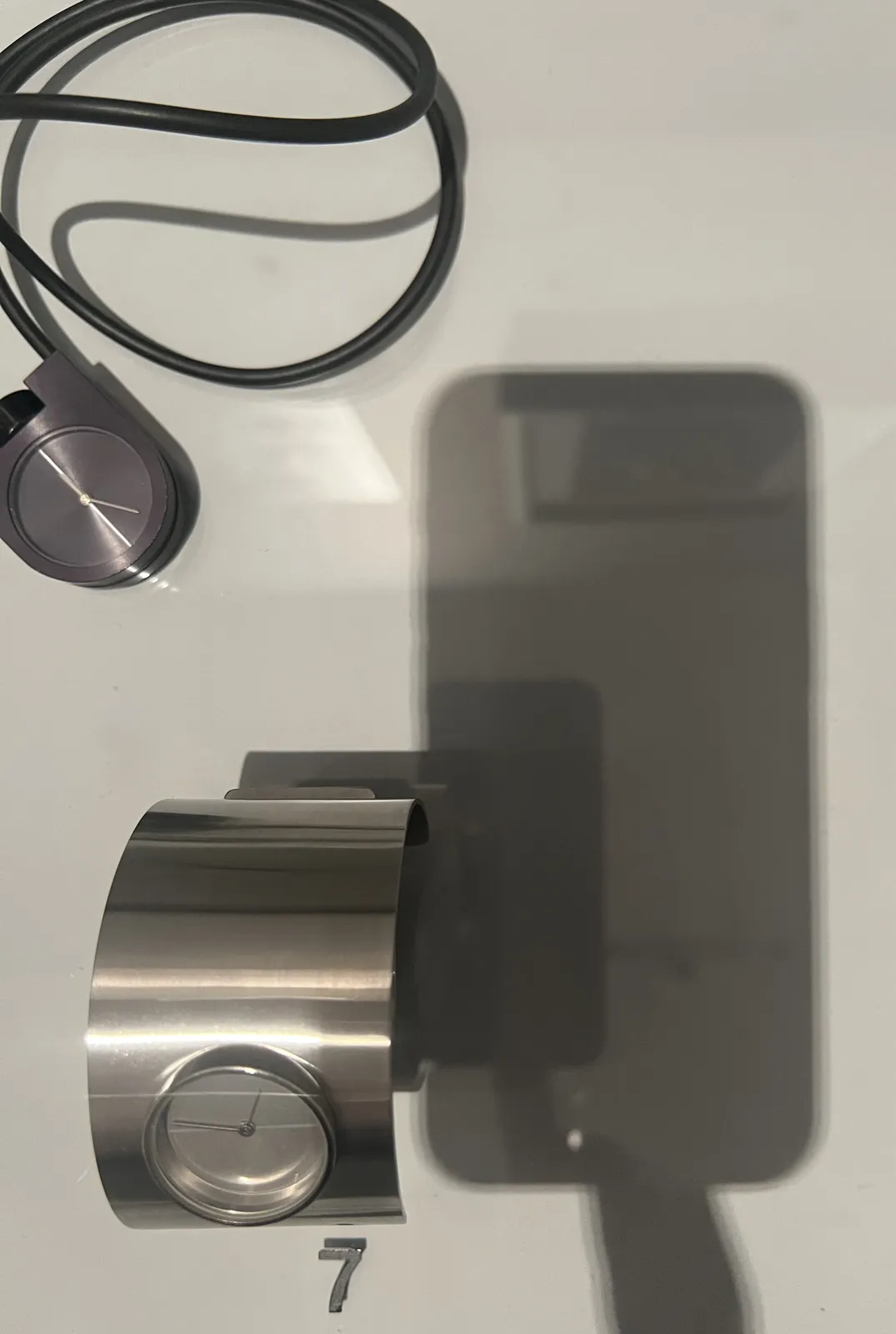 There are two modern looking clocks in this glass showcase. The picture is taken with a phone, which is placed on the glass top, and the photographer is touching the phone with one finger. The shadow of this phone and finger take up the complete right side of the picture. On the left there are two modern, minimalist watches. The one bottom is a wide metal bracelet with a clock in it which seems to point at approximately 14 minutes to one. The one at the top is a necklace, made from a black rubber-like string, and minimalist black and white clock. This clock points at approximately 26 minutes past 11. 