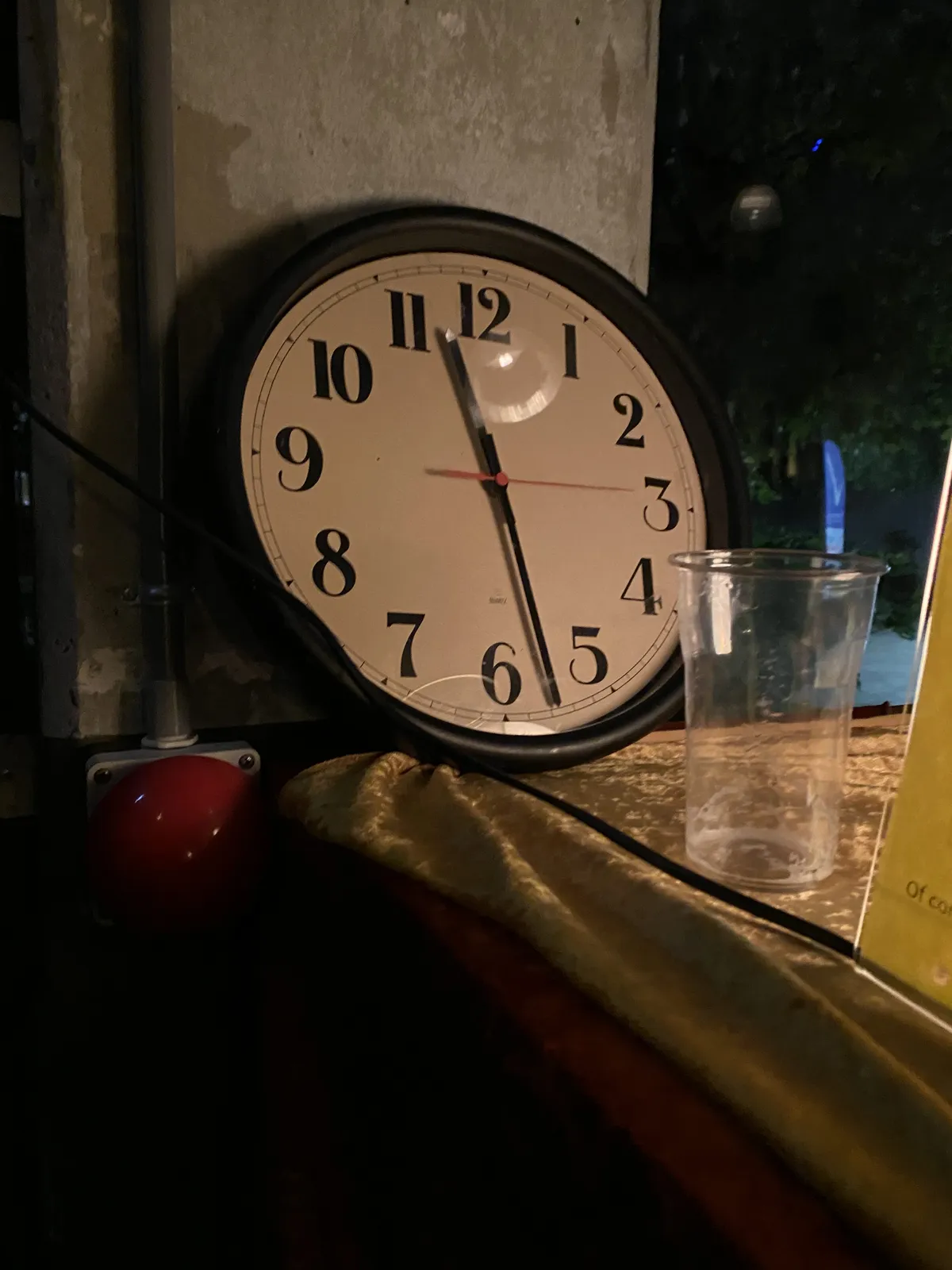A wall clock, leaning against a concrete wall. In front of it is an empty beer glass, next to it is a large, red, emergency button. The clock points at 27 minutes past 11.