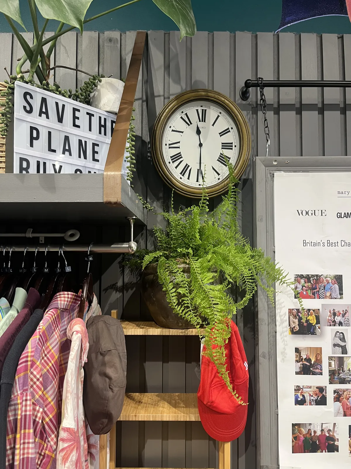 Some shirts hanging on a rack. On top of a shelf there’s a lit sign that says Save The Planet, Shop … The rest of the sign is unreadable. Next to this sign, on the wall, there’s a wall clock with a quasi golden frame, that points at half past eleven.