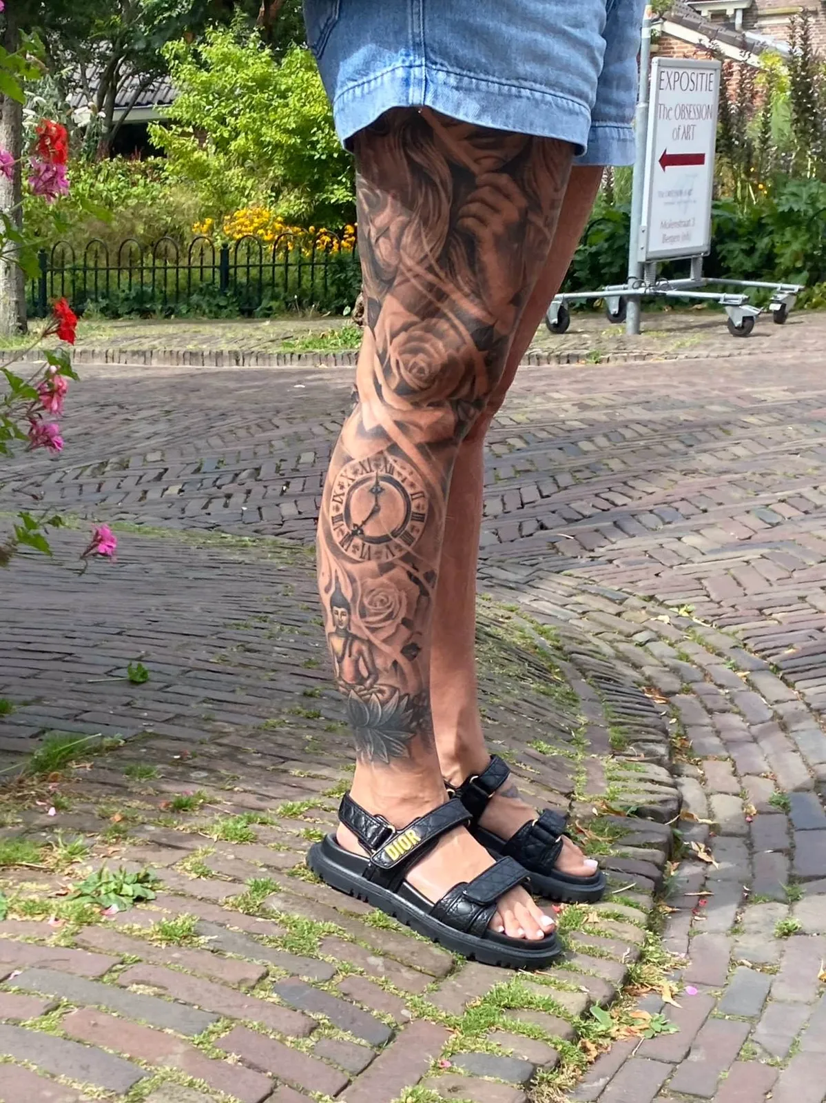 Someone is standing on a sunny, brick lane. Their left leg is covered with a tattoo. There are roses, someone’s face, a buddha, and a clock in this tattoo. The clock points at 25 minutes to 12.