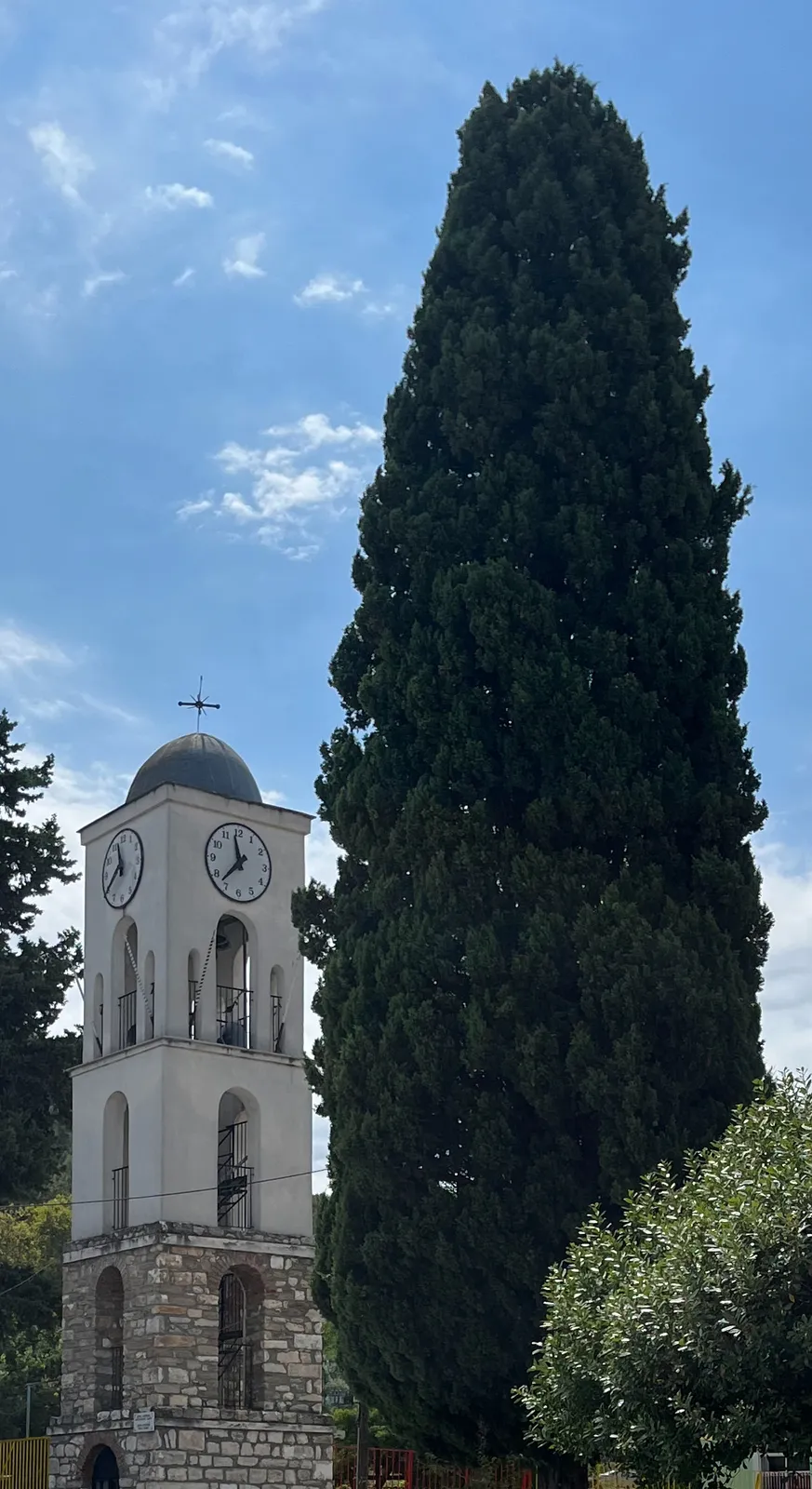 A huge cypress tree that dwarves this church tower in old Thasos town. On the tower there’s a clock that points at 23 minutes to 12.
