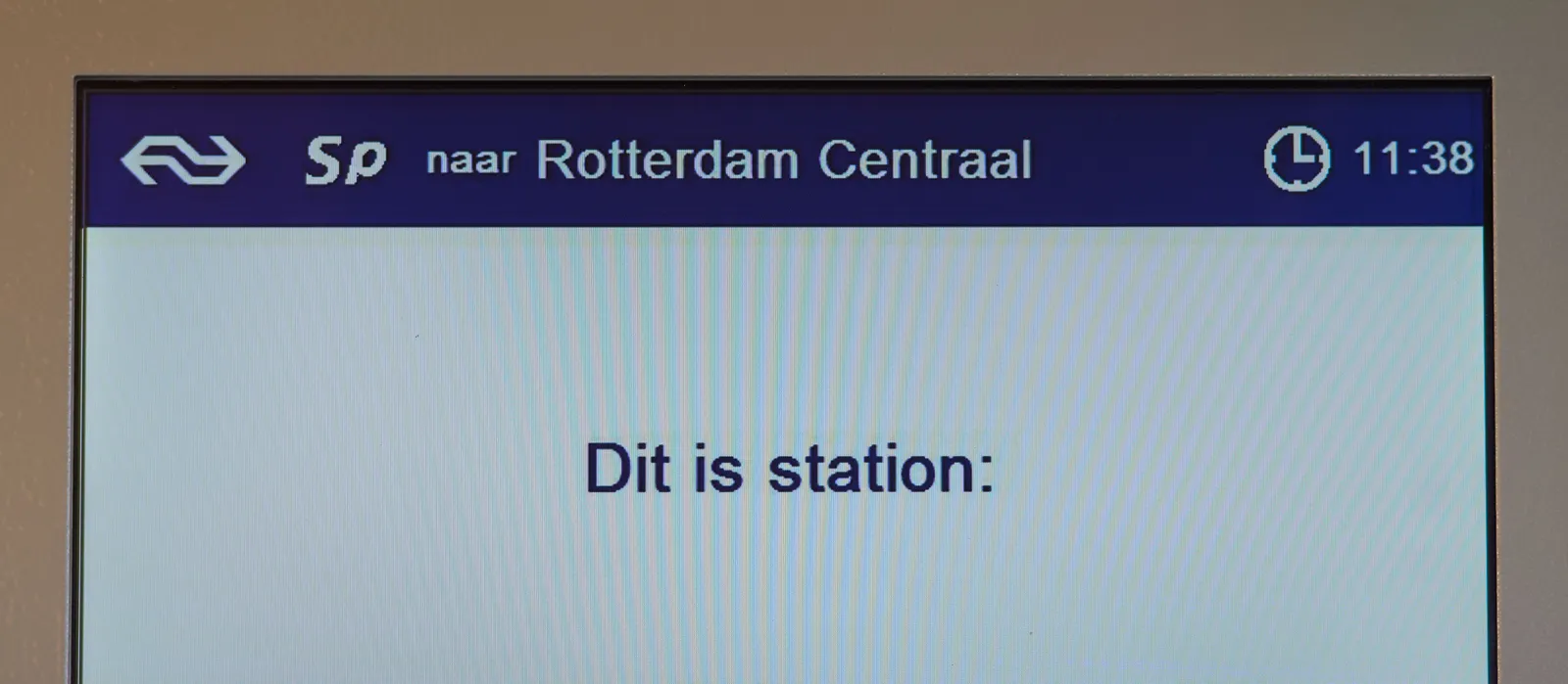 A display in a train says this is a train to Rotterdam. It also shows the words, translated from Dutch, this is station, but the name of what station this is are outside the picture frame. The time is shown in the top right corner of this display and it says it’s 11:38. 