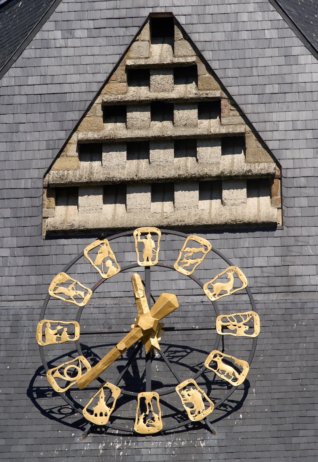 A large, shiny golden clock on the black roof of a church. The hour indicarors are made from golden icons, depicting country life, and more specifically, the hunt. The enormous golden hands point at 22 minutes to twelve. 