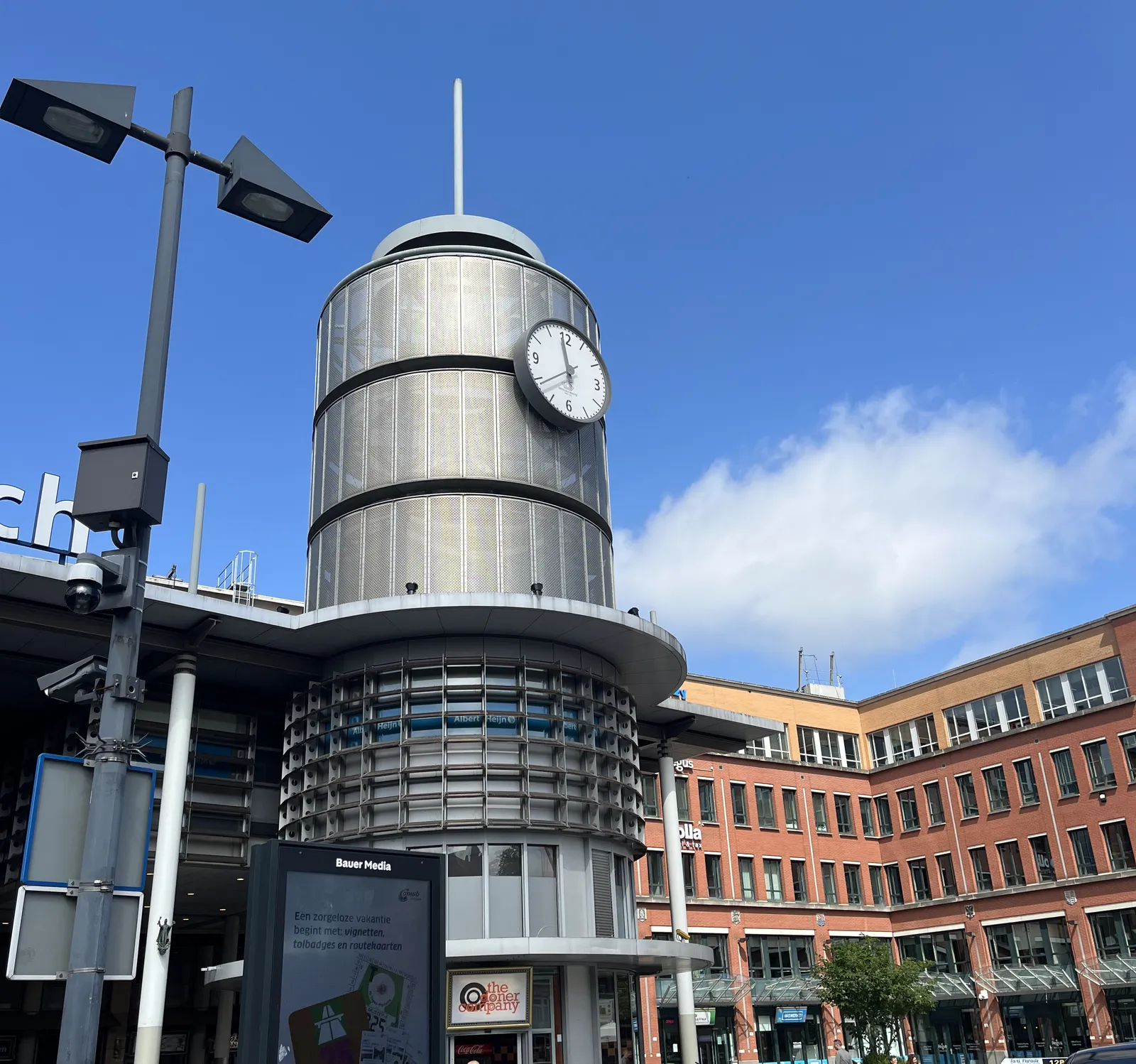 A post modernist brick building, with a large cylindric tower made from some sort of metal, or plastic. On this tower is a clock that points at 22 minutes to twelve. 