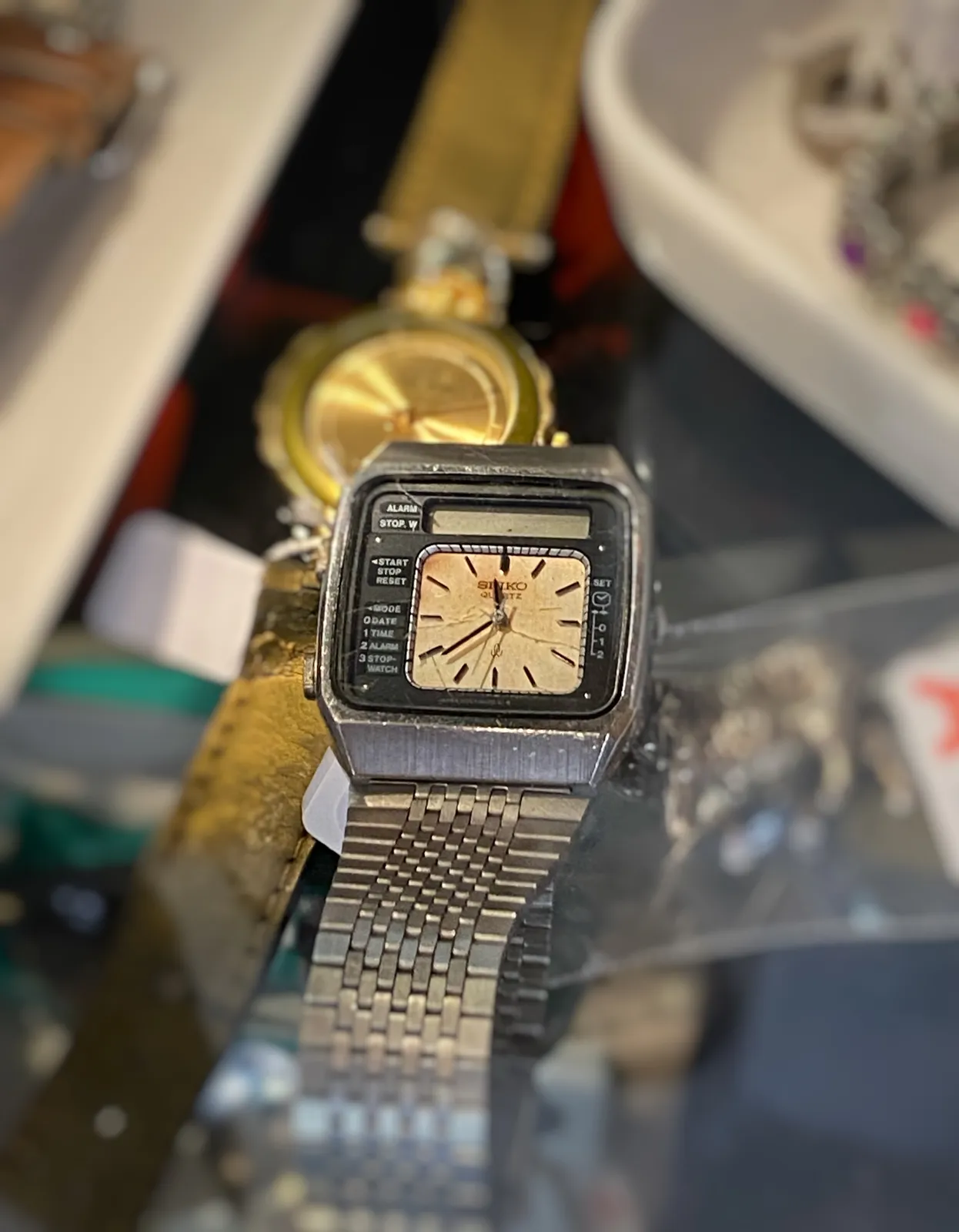 A square wrist watch. In a smaller square the time is shown. Around it there are all kinds of words printed: Alarm stop, start stop reset, mode, 0 date 1 time 2 alarm 3 stop watch. The time on this clock is 21 minutes to 12.