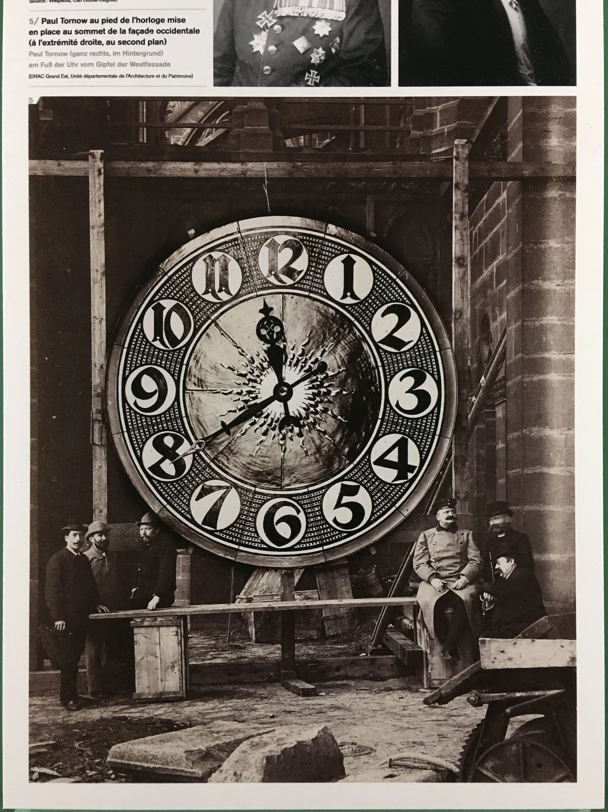 Six men with impressive moustaches and beards, all wearing hats, on this black and white image. They stand next to an enormous clock which points at 20 minutes to 12.