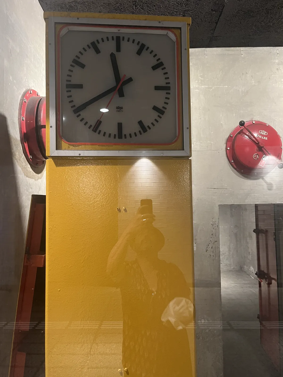 A modernist clock, on a yellow column. In the background, on bare concrete, there are two, large, red alarm bells. The clock points at 20 to twelve.