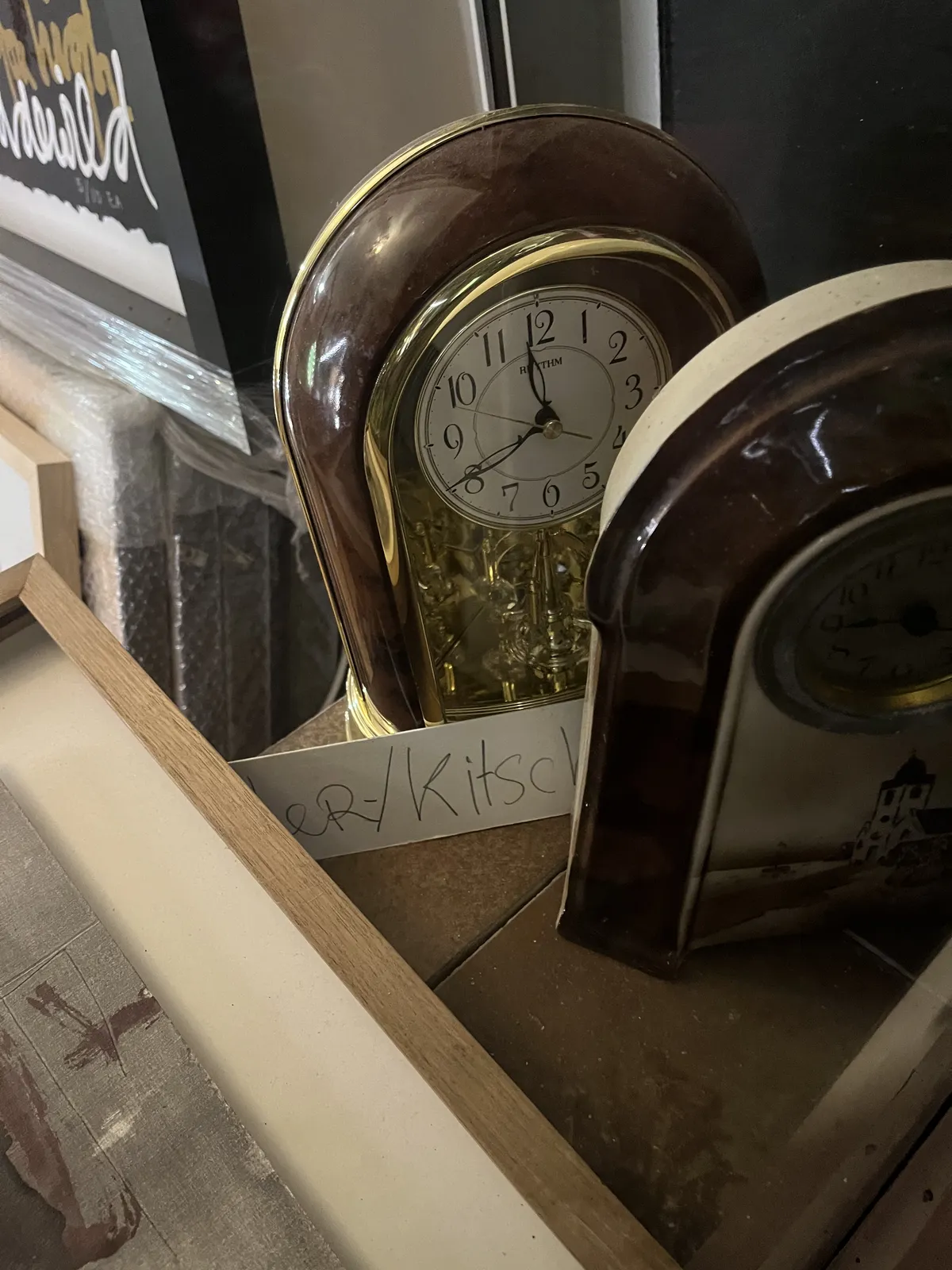 A small table clock standing behind another small table clock. There’s a sign with the handwritten word ‘kitsch’ on it. This small clock points at 20 minutes to twelve.
