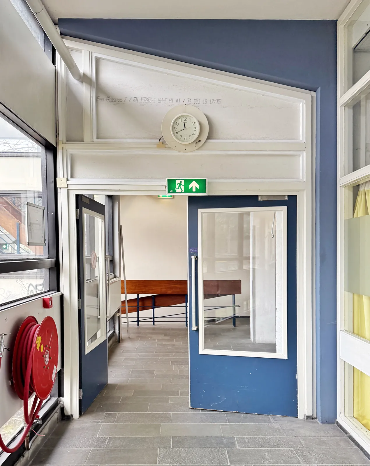 Above a blue double door, in a hallway, there’s a clock that points at 19 minutes to twelve.