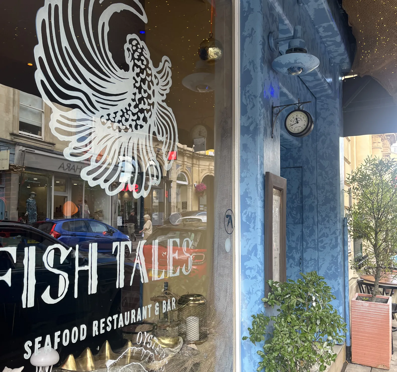 On the window it says that this is Fish Tales, a seafood restaurant. Attached to the facade of this restaurant is a small clock which points at 18 minutes to twelve. 