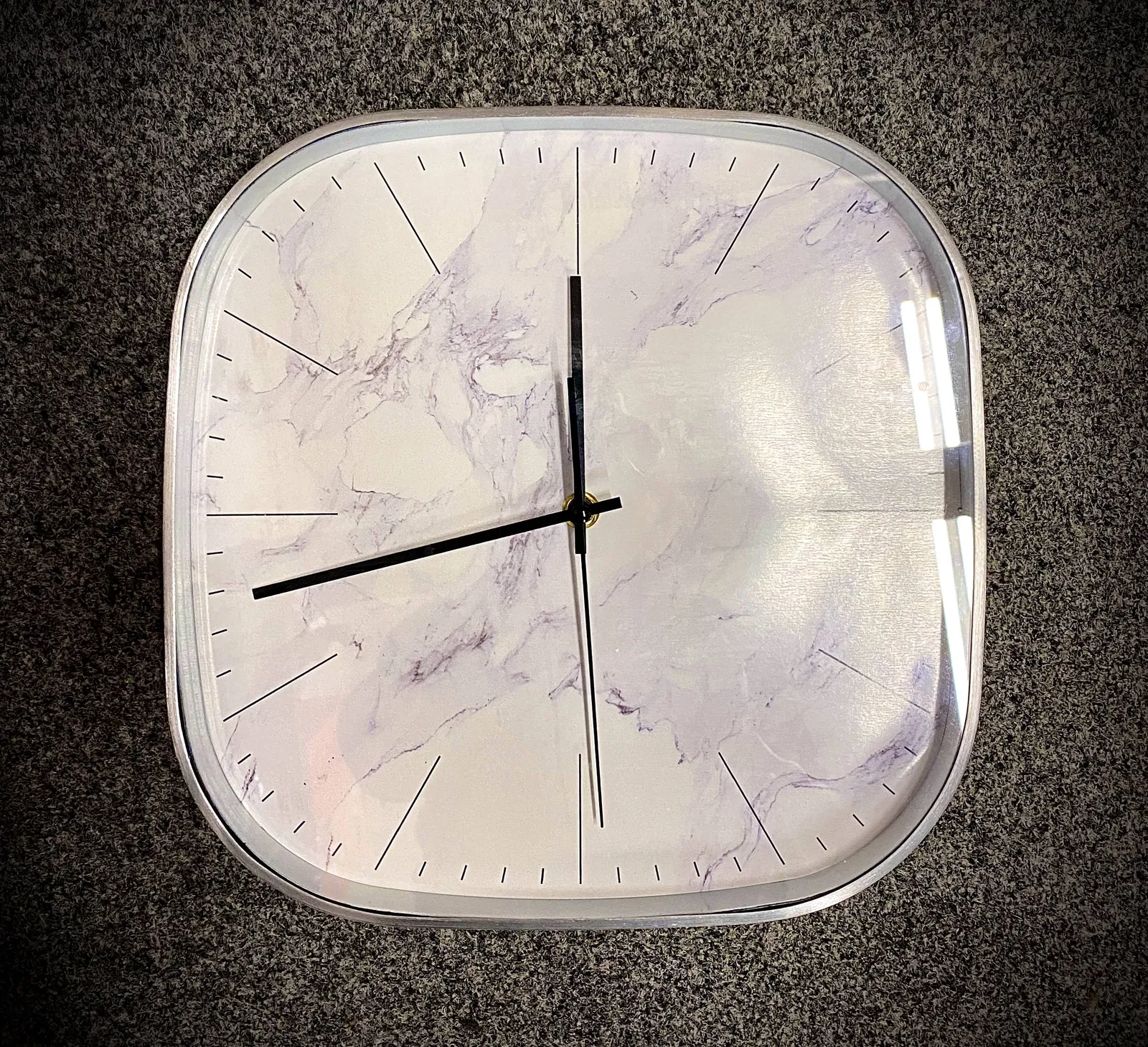 A simple, square wall clock with rounded corners. It points at 18 minutes to 12.