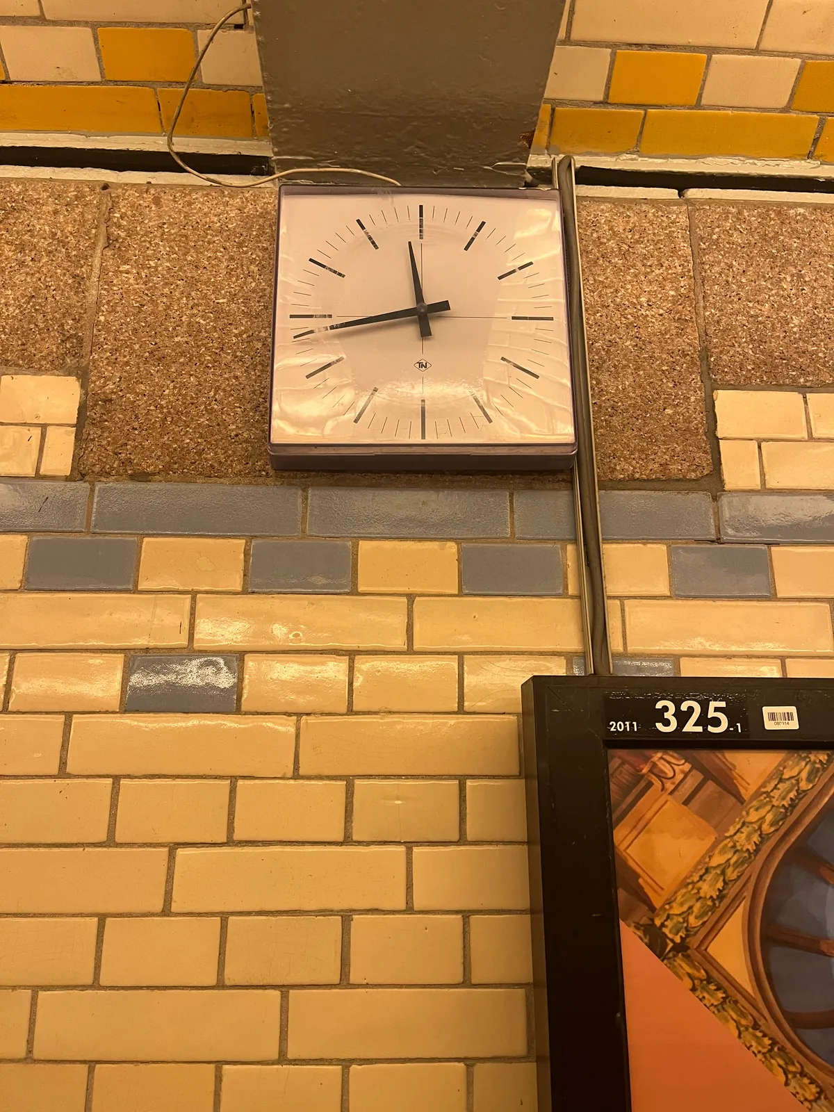 A part of an advertising billboard on a wall made from colourful tiles. There’s a square clock with pointy hands and thin, long indicators. It points at 17 minutes to 12.