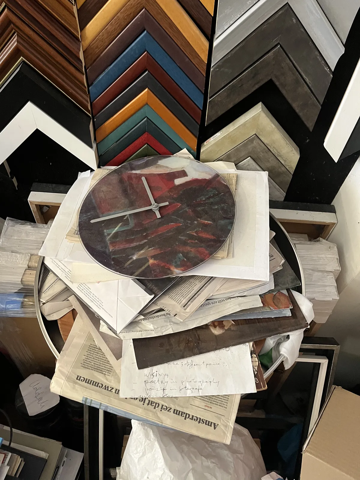 On a pile of old newspapers, in front of examples of different types of art frames, there’s a clock whise face seems to be made from red marble. There are no indicators at all on this clock. The two hands form an exact 90 degree angle, so since there are no indicators it could be many different timestamps. But by assuming that the clock is laid down orderly, with the twelve pointing up, we can conclude that it is about 16 minutes to twelve 