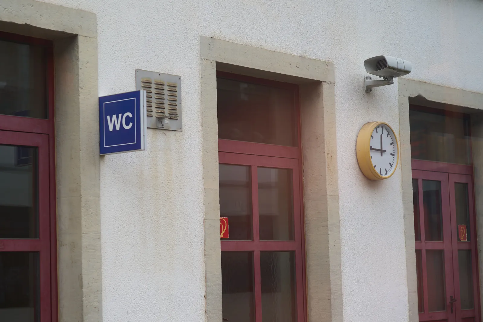 There are three doors in this white building. Next to one of them there’s a sing that says WC. Next to one of the other doors there’s a clock that points at 16 minutes to 12.